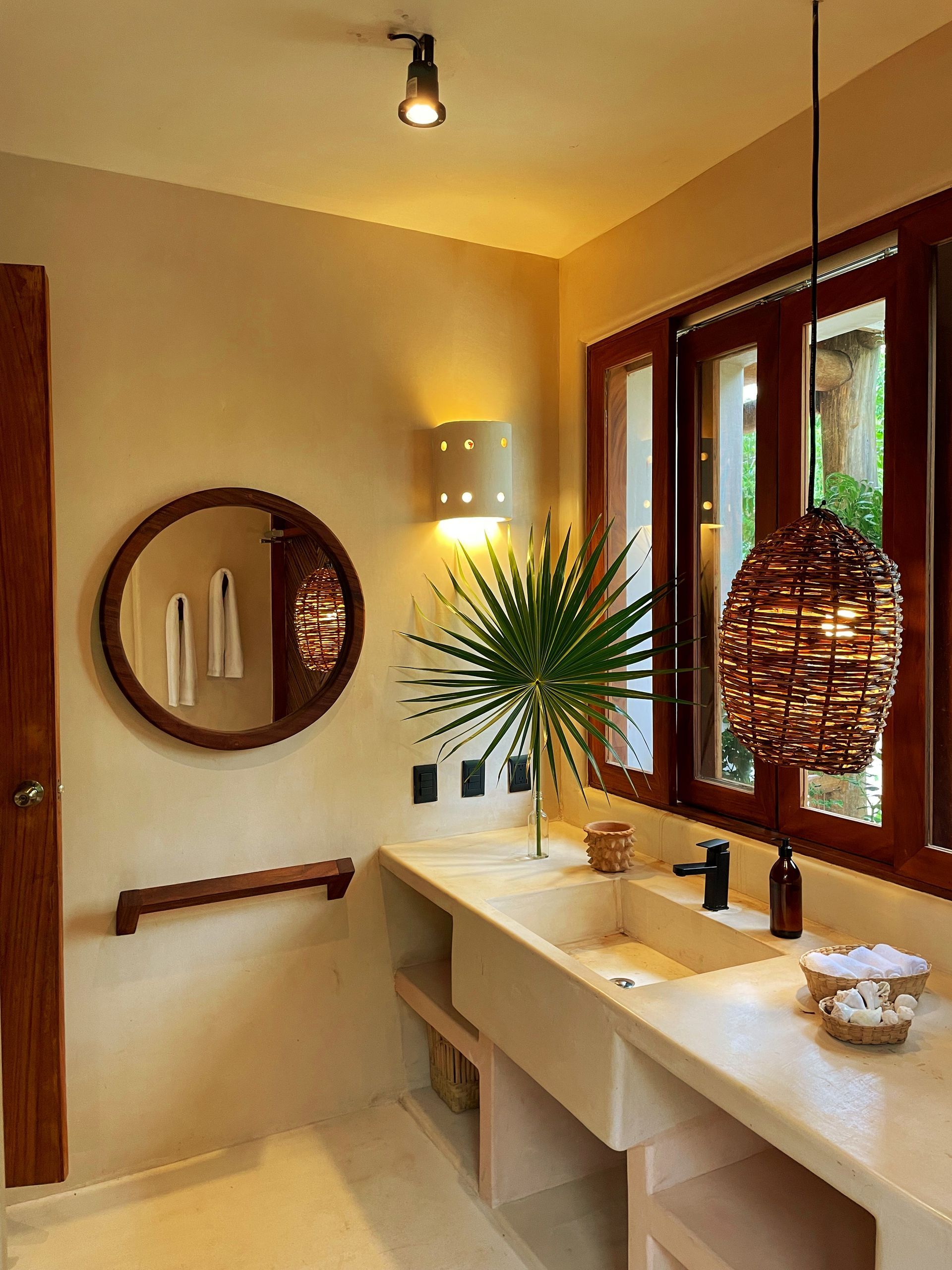 Baño con decoración rústica, espejo redondo, lámpara colgante de mimbre y lavabo blanco.