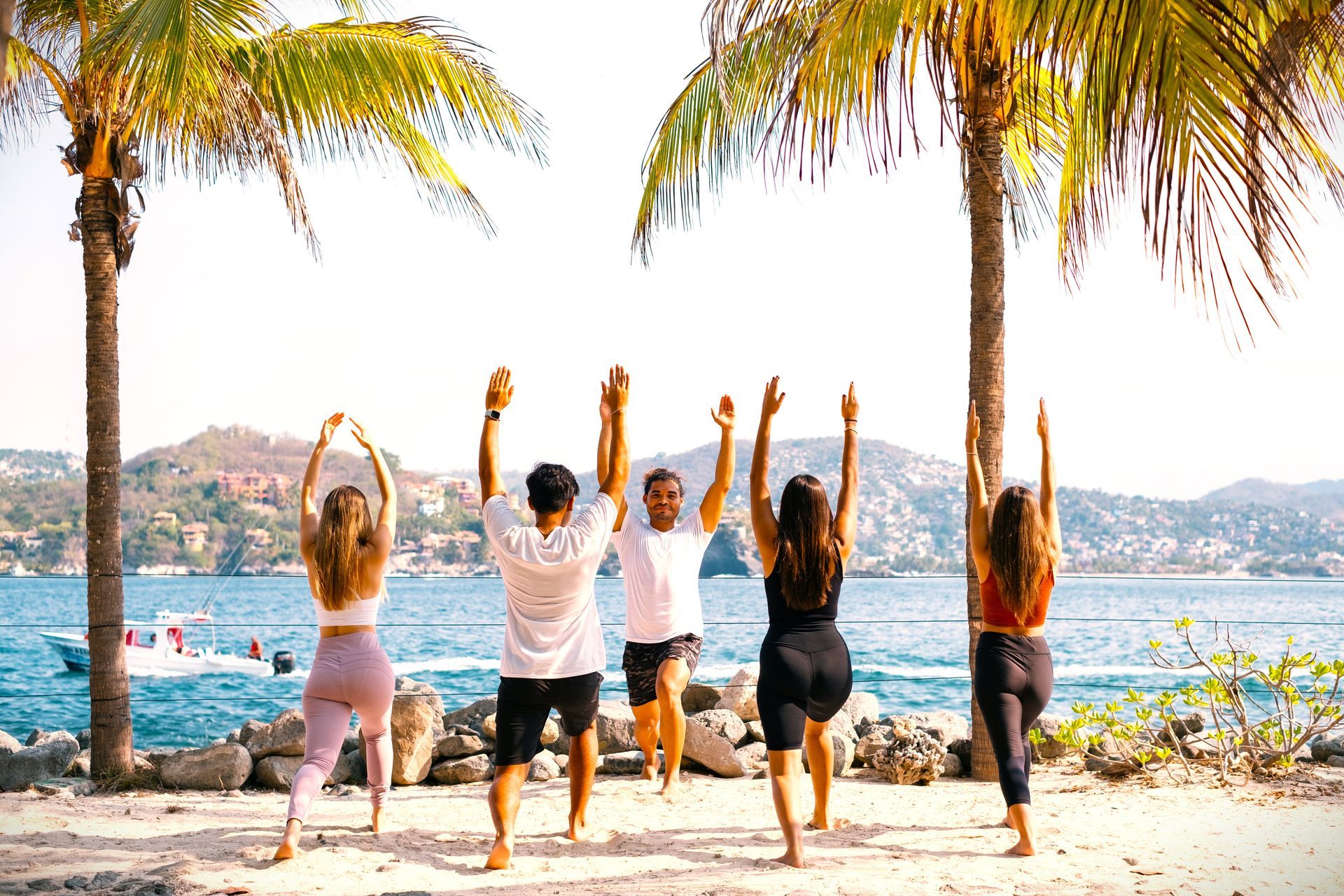 Personas practicando yoga en una playa bajo palmeras, con los brazos en alto. Montañas y agua al fondo.