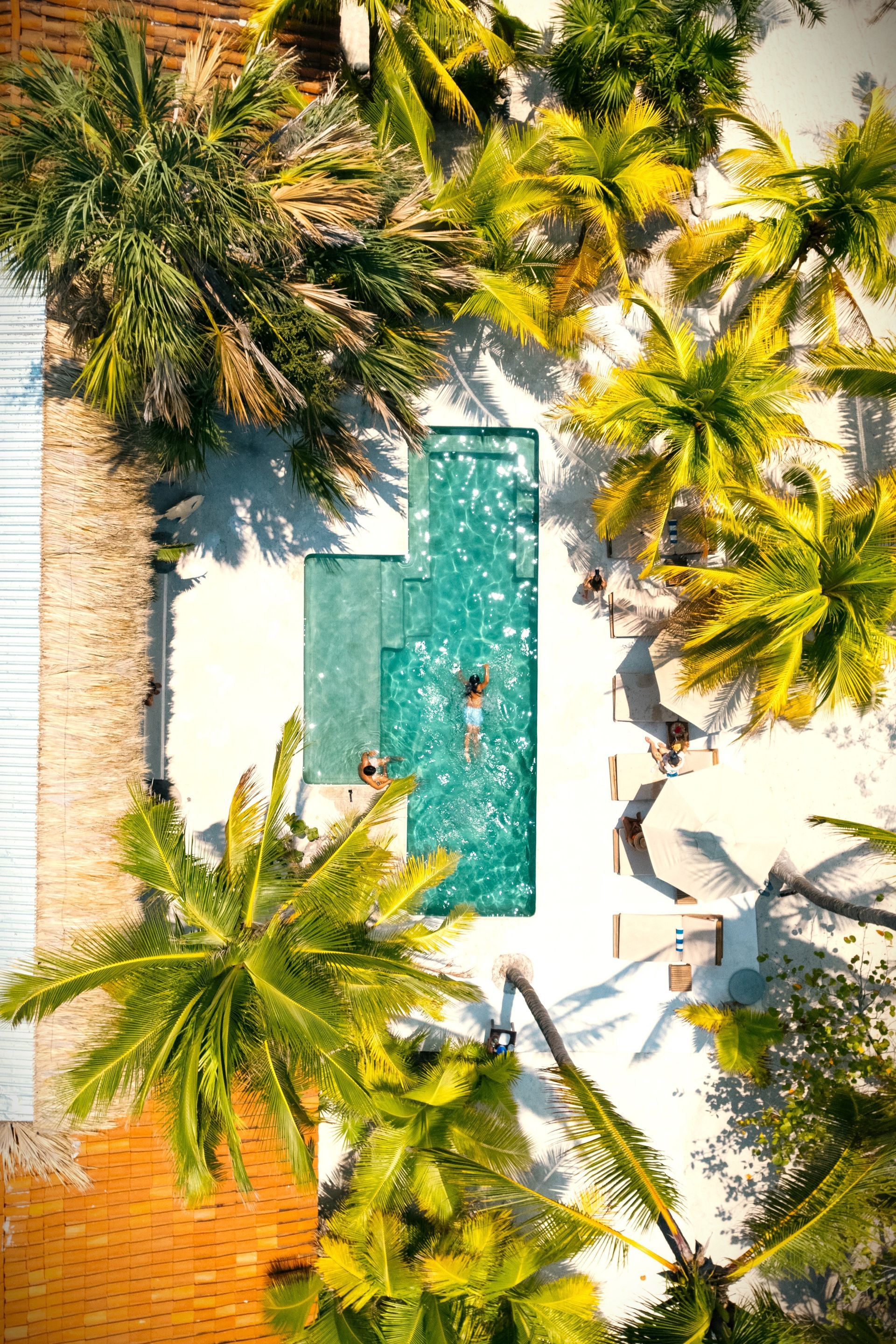 Vista aérea de una piscina turquesa con gente, rodeada de palmeras y arena blanca, cerca de un edificio de ladrillo.