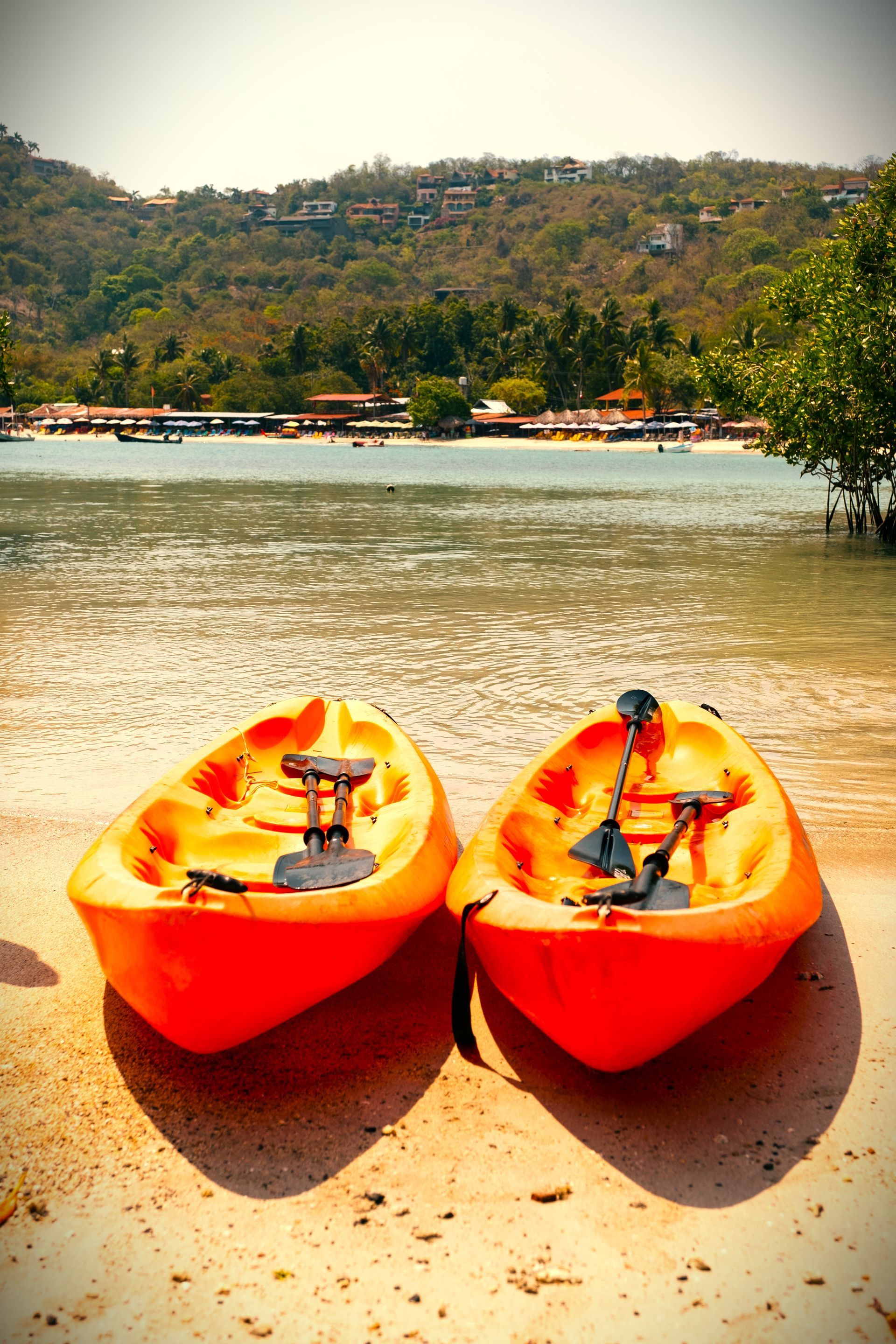 Dos kayaks naranjas en una playa de arena con agua y una ladera cubierta de árboles al fondo.