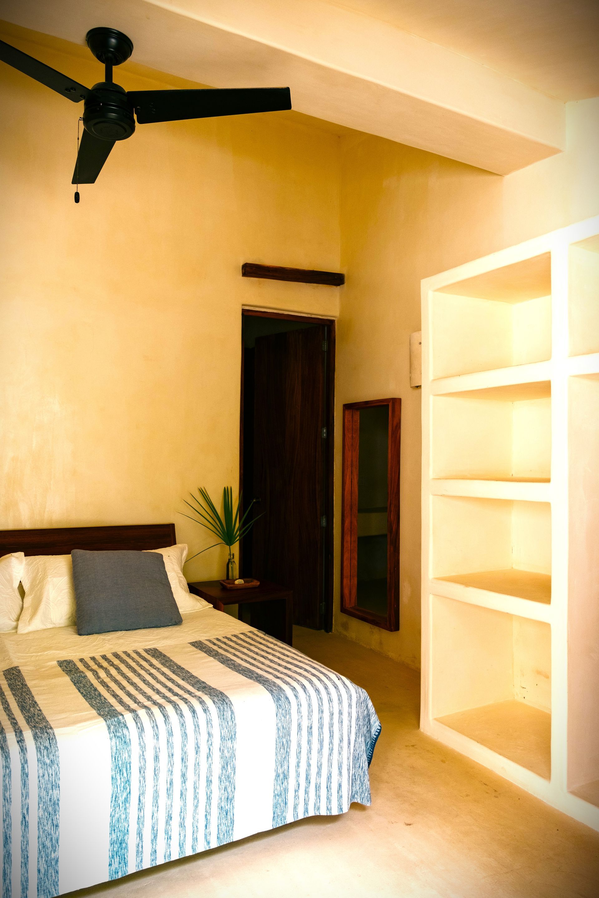 Dormitorio con cama, estantería empotrada, ventilador de techo oscuro y puerta. Paredes de color amarillo pálido.