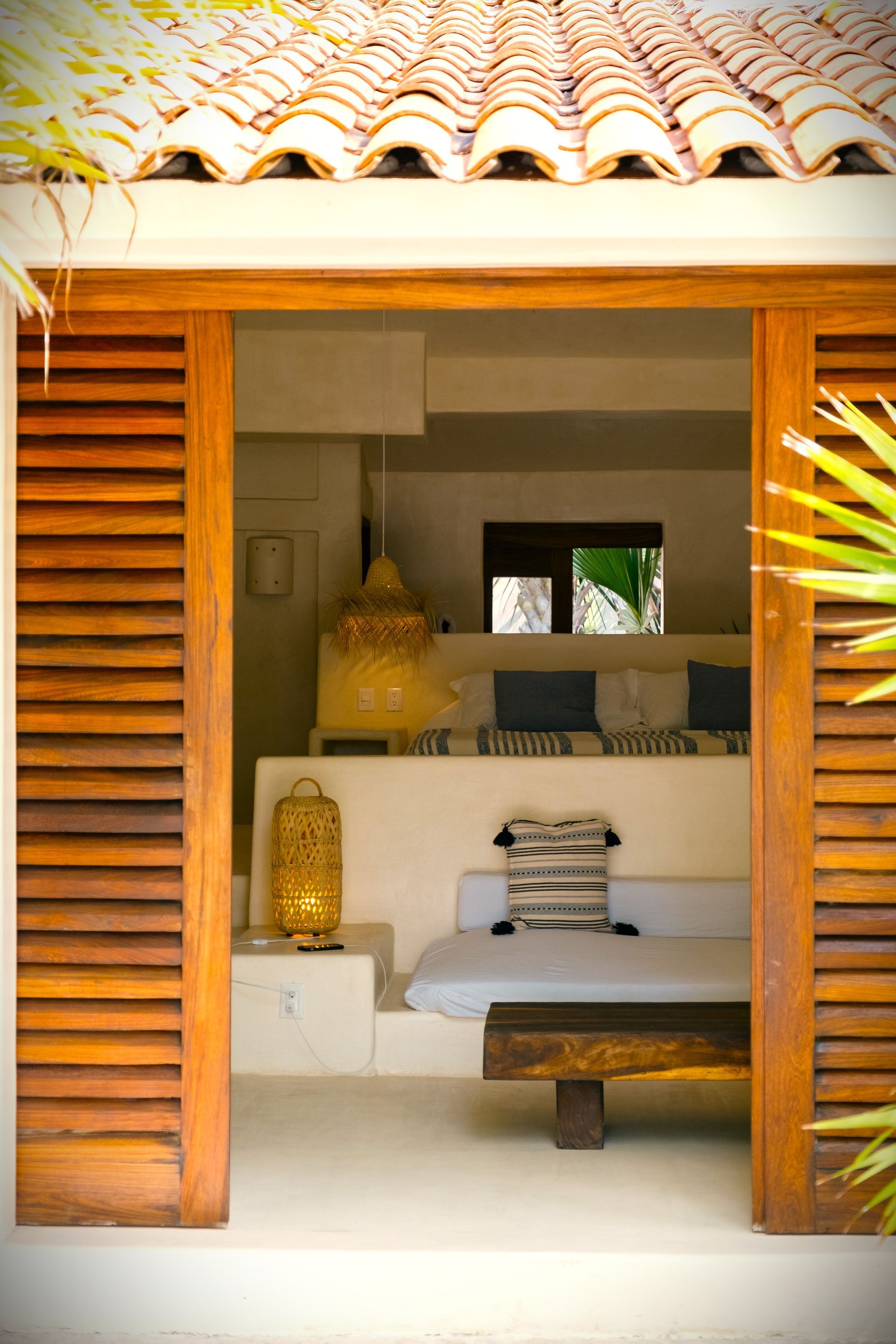 Una puerta con contraventanas de madera enmarca un interior de paredes blancas con un sofá, un banco e iluminación. 