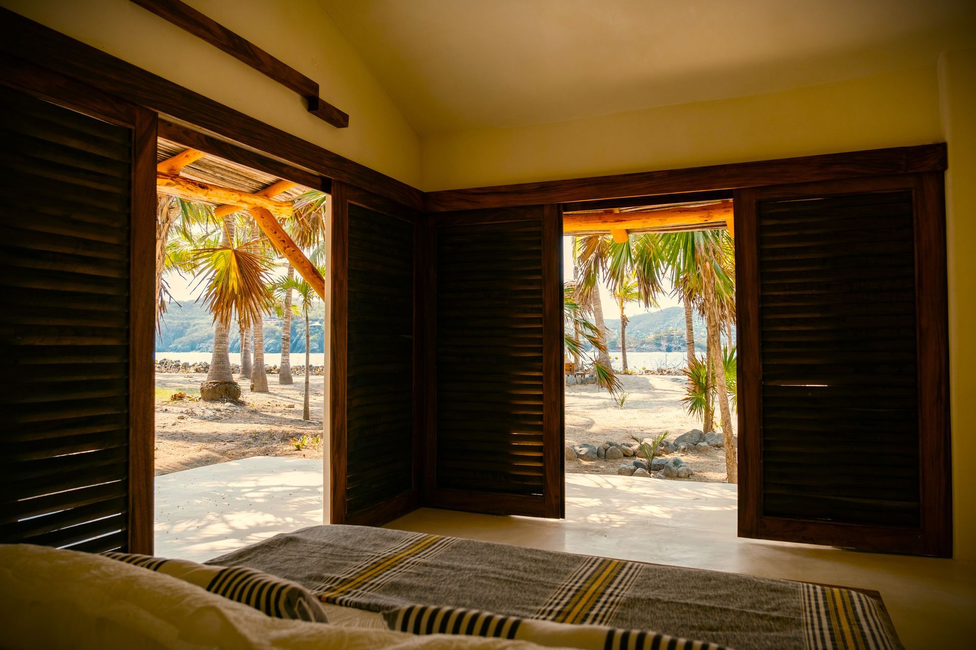 Dormitorio con puertas abiertas que dan a una playa con palmeras y agua, y con luz solar brillando a través de ella.