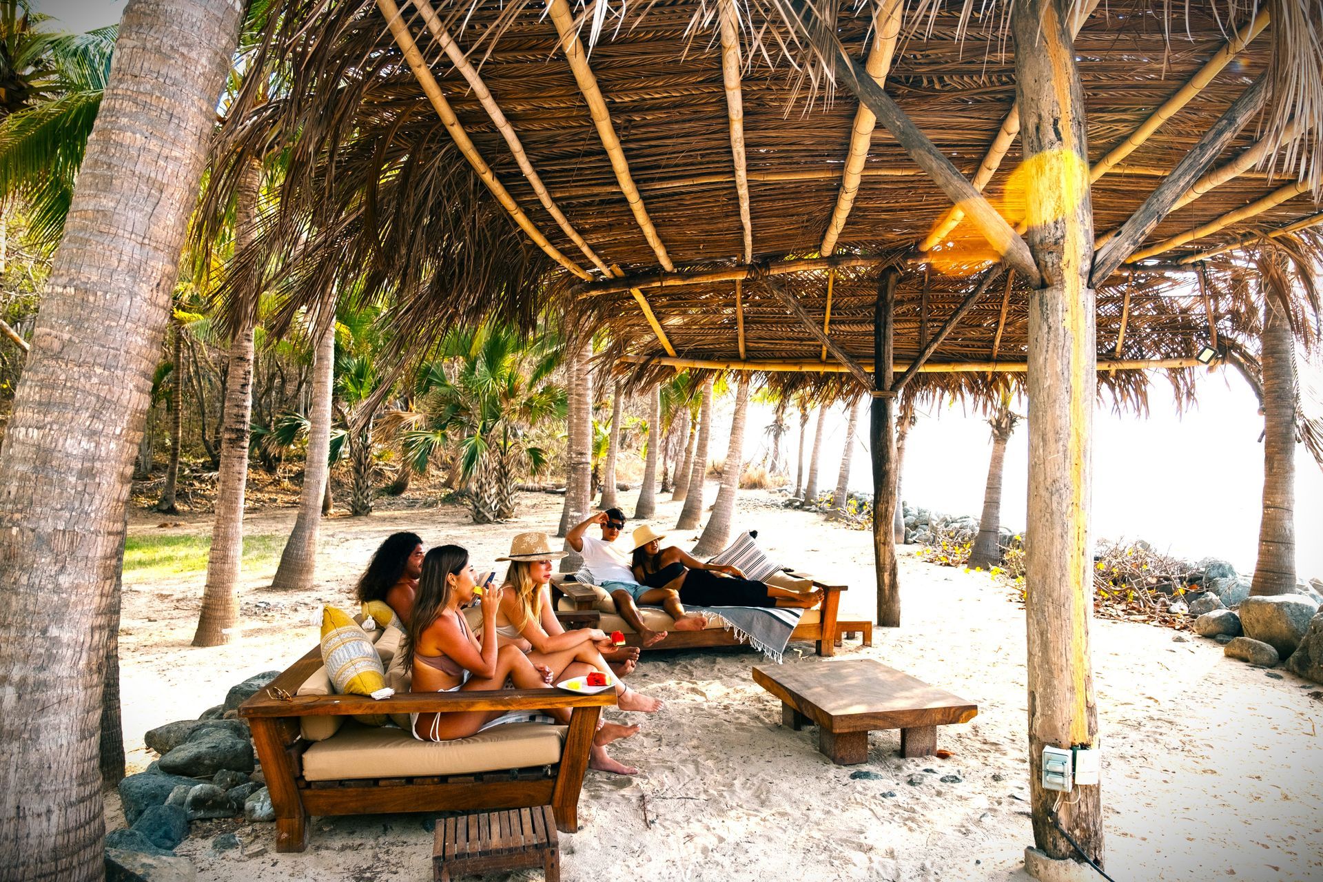Gente relajándose en tumbonas de playa bajo una estructura con techo de paja en una playa de arena.