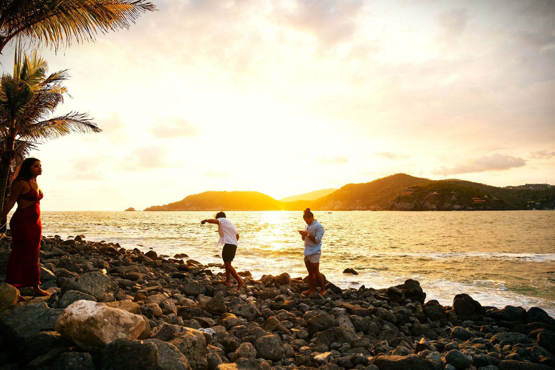 Escena de atardecer en una playa rocosa con tres personas; palmera, agua y colinas.