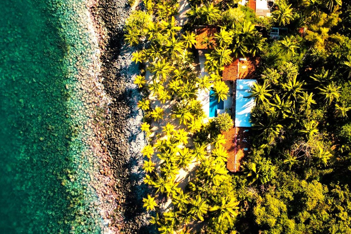 Vista aérea: agua turquesa, orilla rocosa, exuberantes árboles verdes que rodean un edificio con una piscina azul.