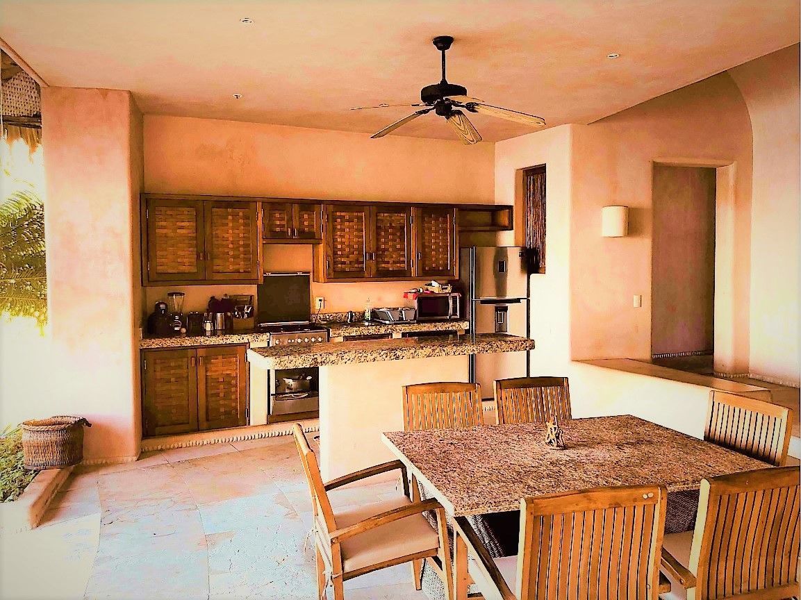 Cocina exterior con gabinetes de madera y encimera de granito. Mesa de comedor y sillas en primer plano.