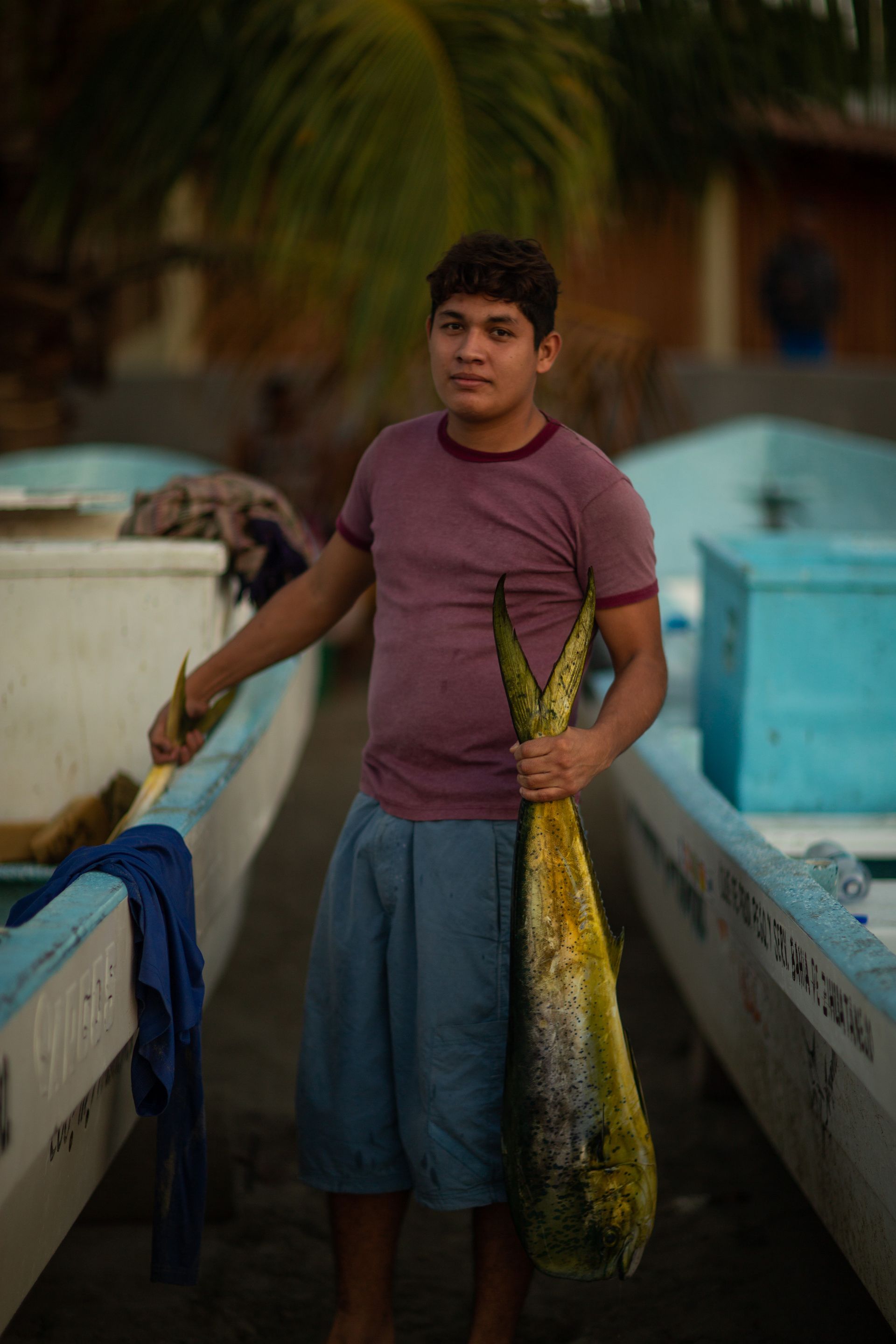 Un joven sostiene un pez grande, de pie entre dos botes. Lleva una camisa granate y pantalones cortos azules.