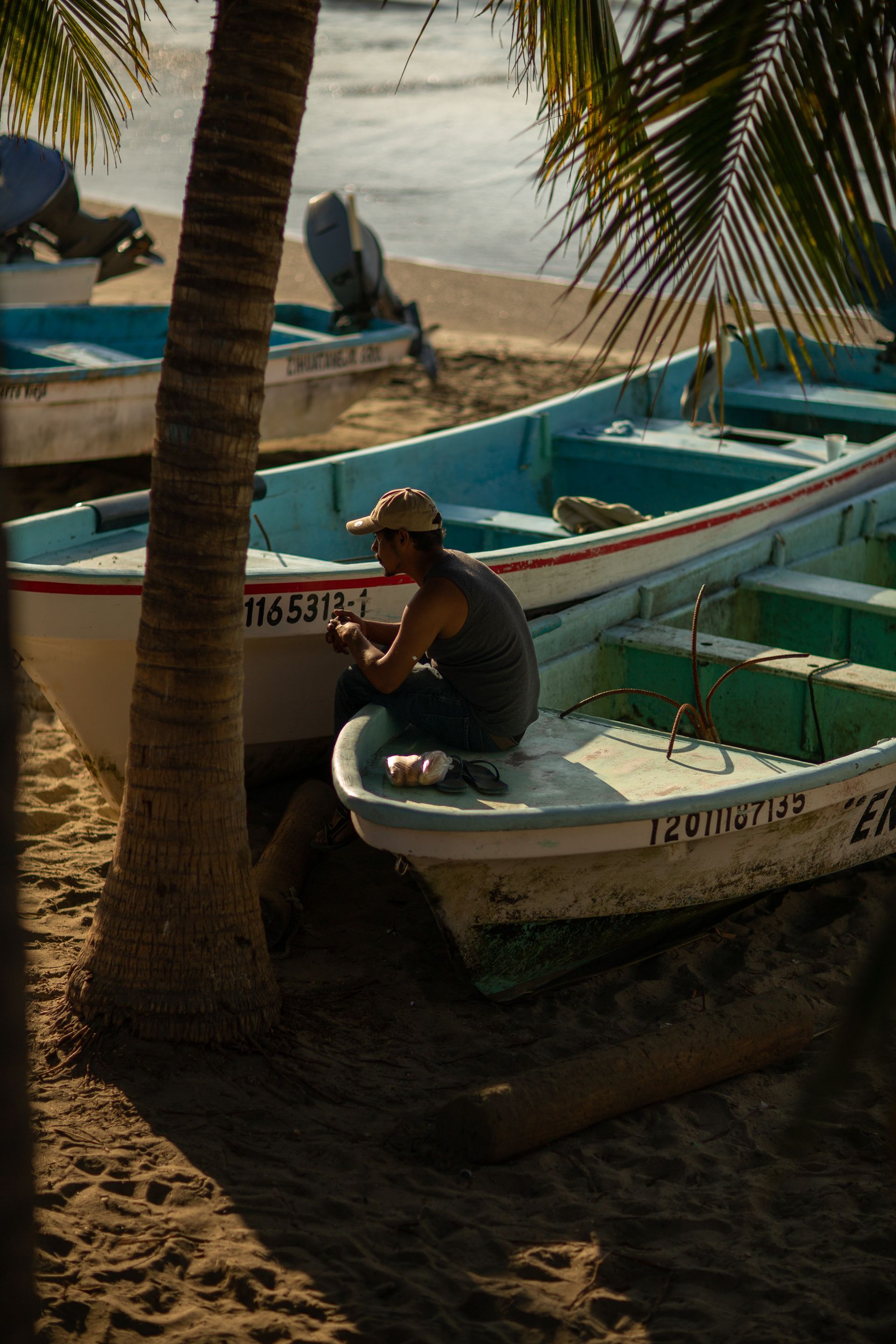 Un hombre trabaja en un pequeño bote en una playa de arena. Barcos, palmeras y el océano al fondo.