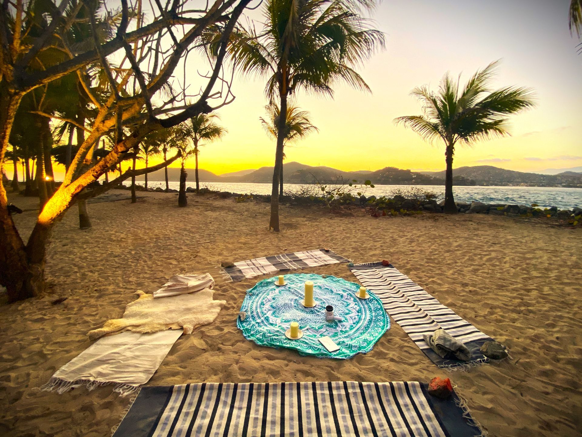 Instalación de playa para yoga o meditación al atardecer; colchonetas, velas, vista al mar, palmeras.