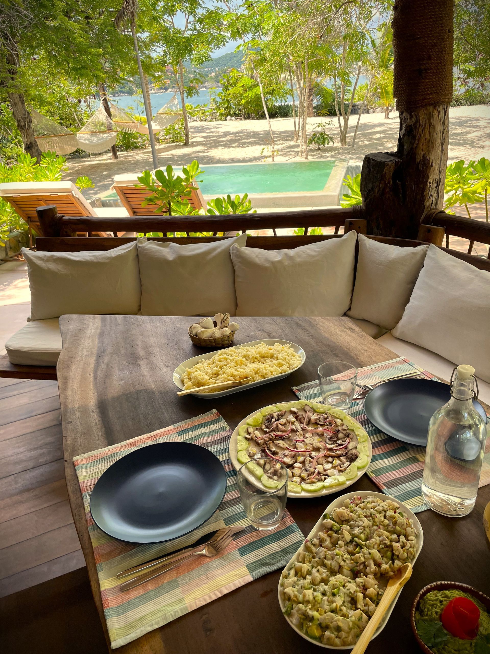 Mesa de comedor al aire libre con comida y platos, con vista a la playa y la piscina.