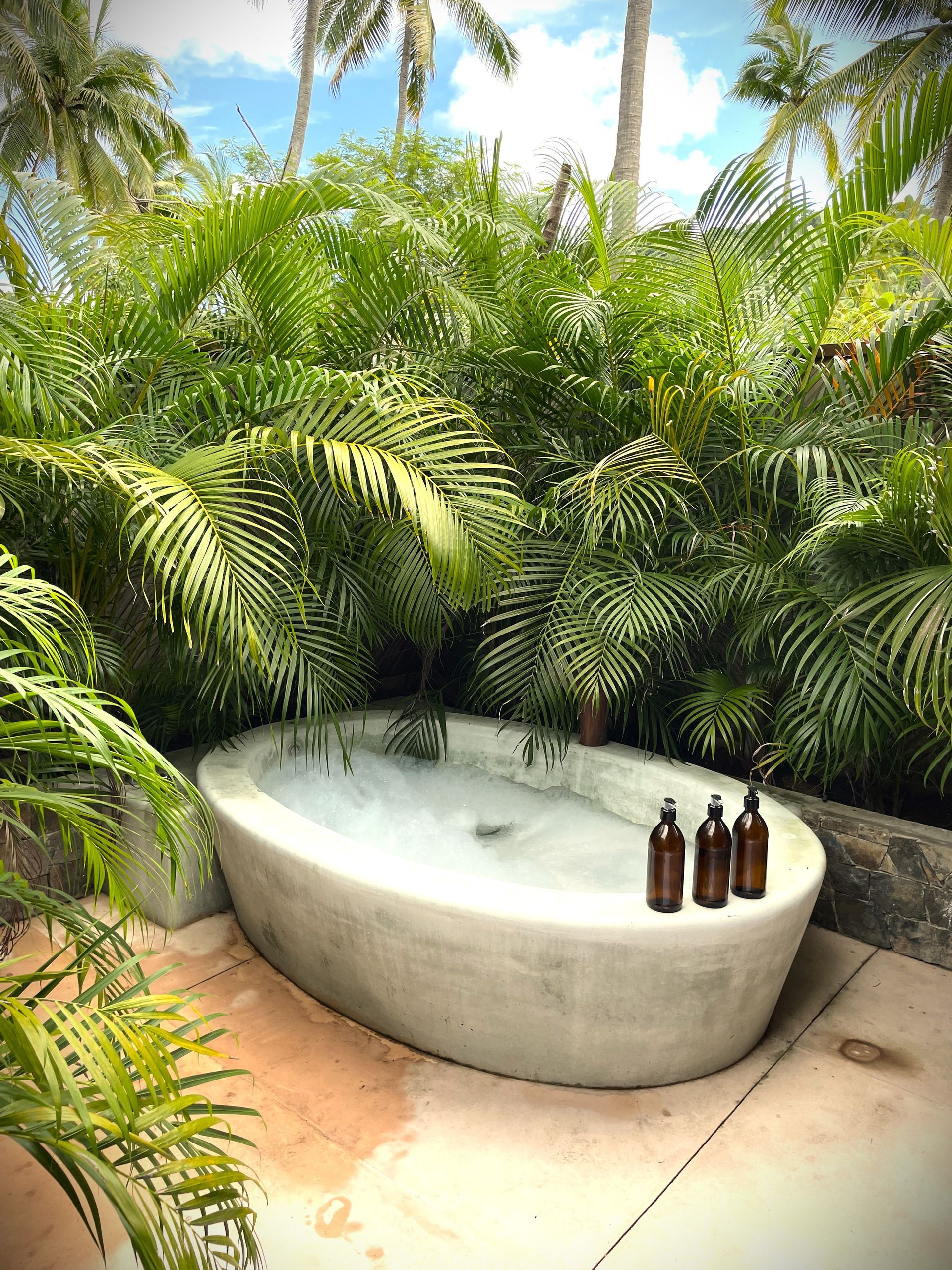 Bañera de piedra rodeada de exuberante vegetación, con baño de burbujas y tres botellas marrones.