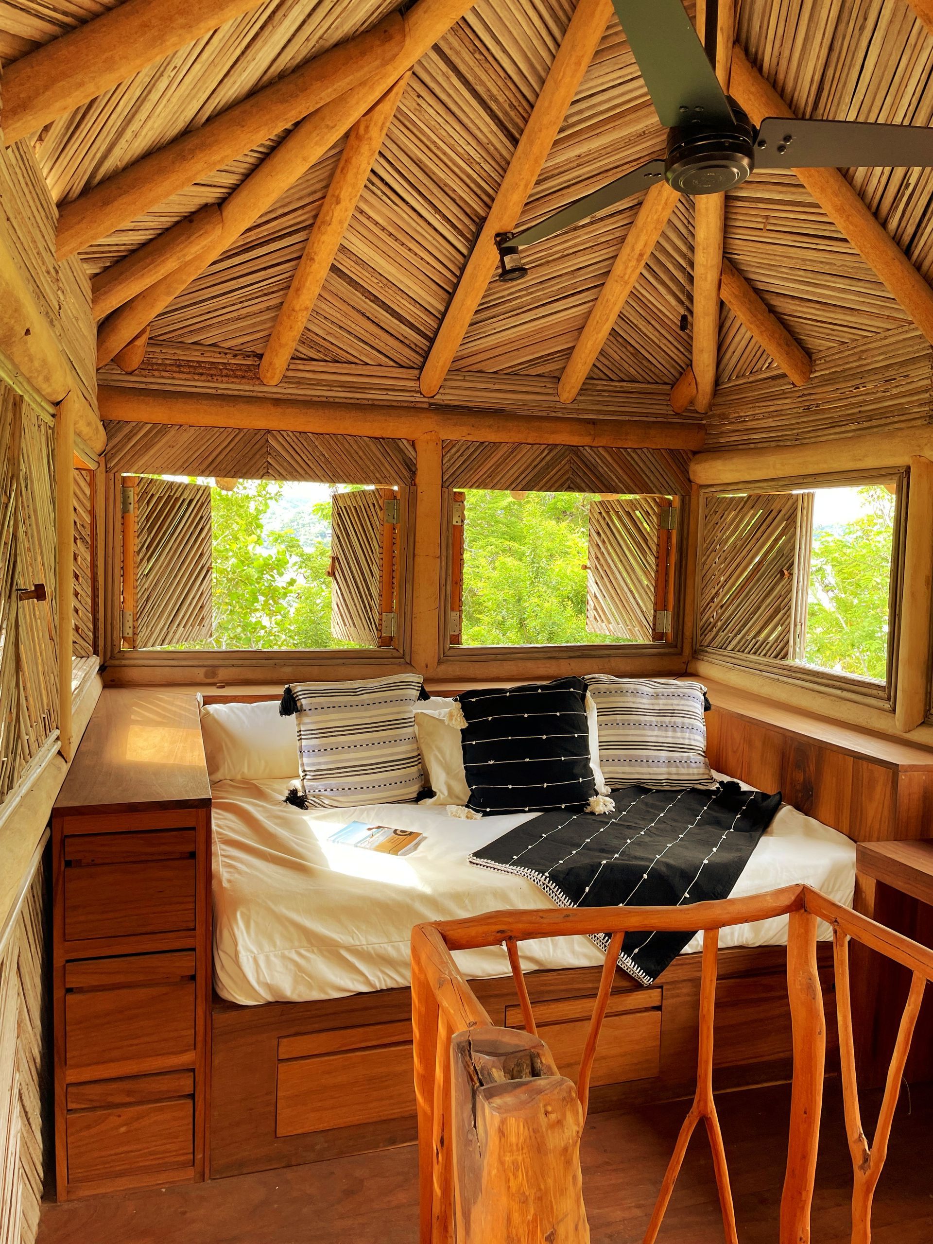 Acogedor dormitorio en una casa del árbol de madera con cama, ventanas y techo de paja, rodeado de árboles verdes.