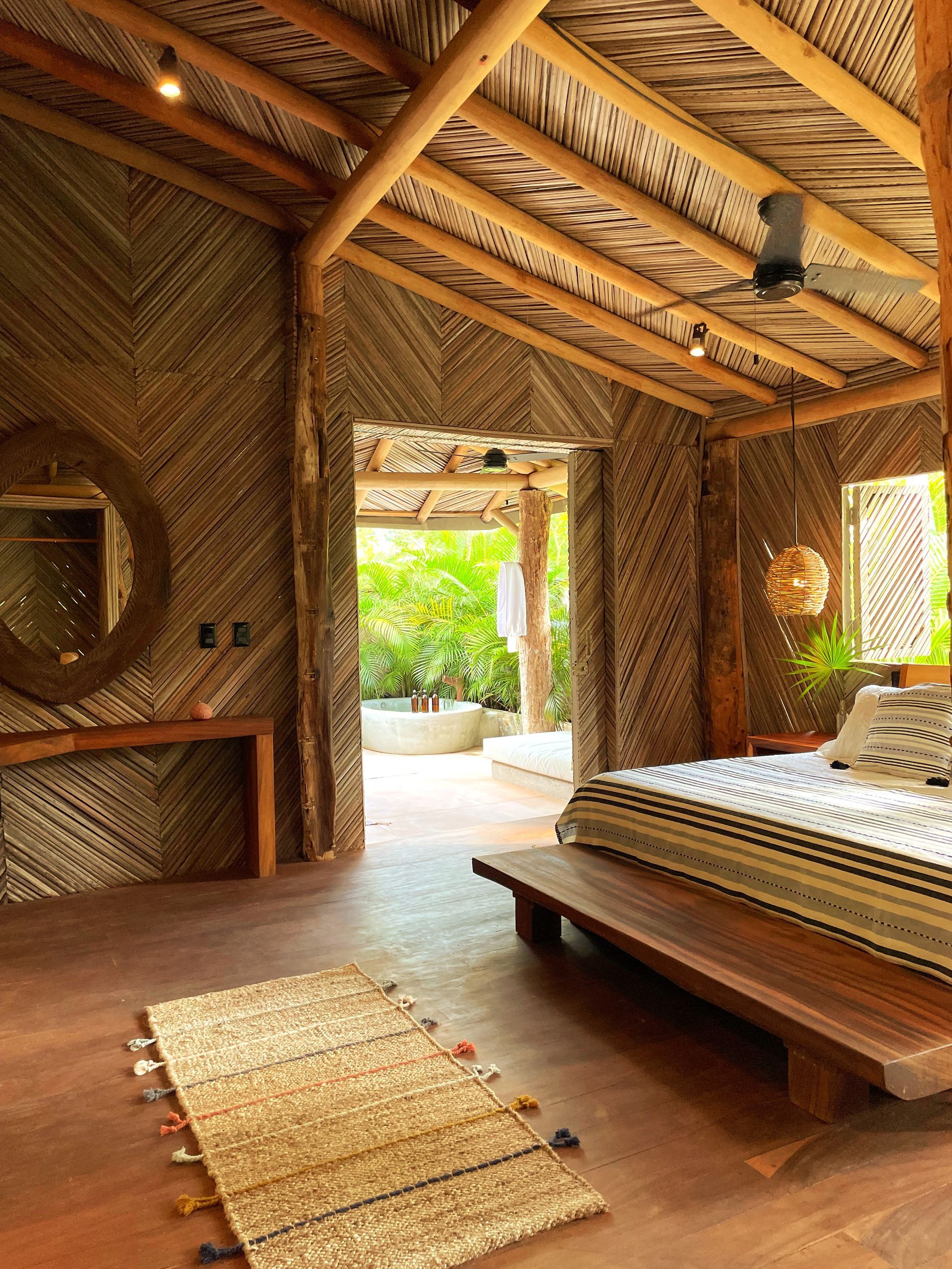 Dormitorio con detalles en madera, paredes y techo de hojas de palma, puerta abierta a la vegetación.