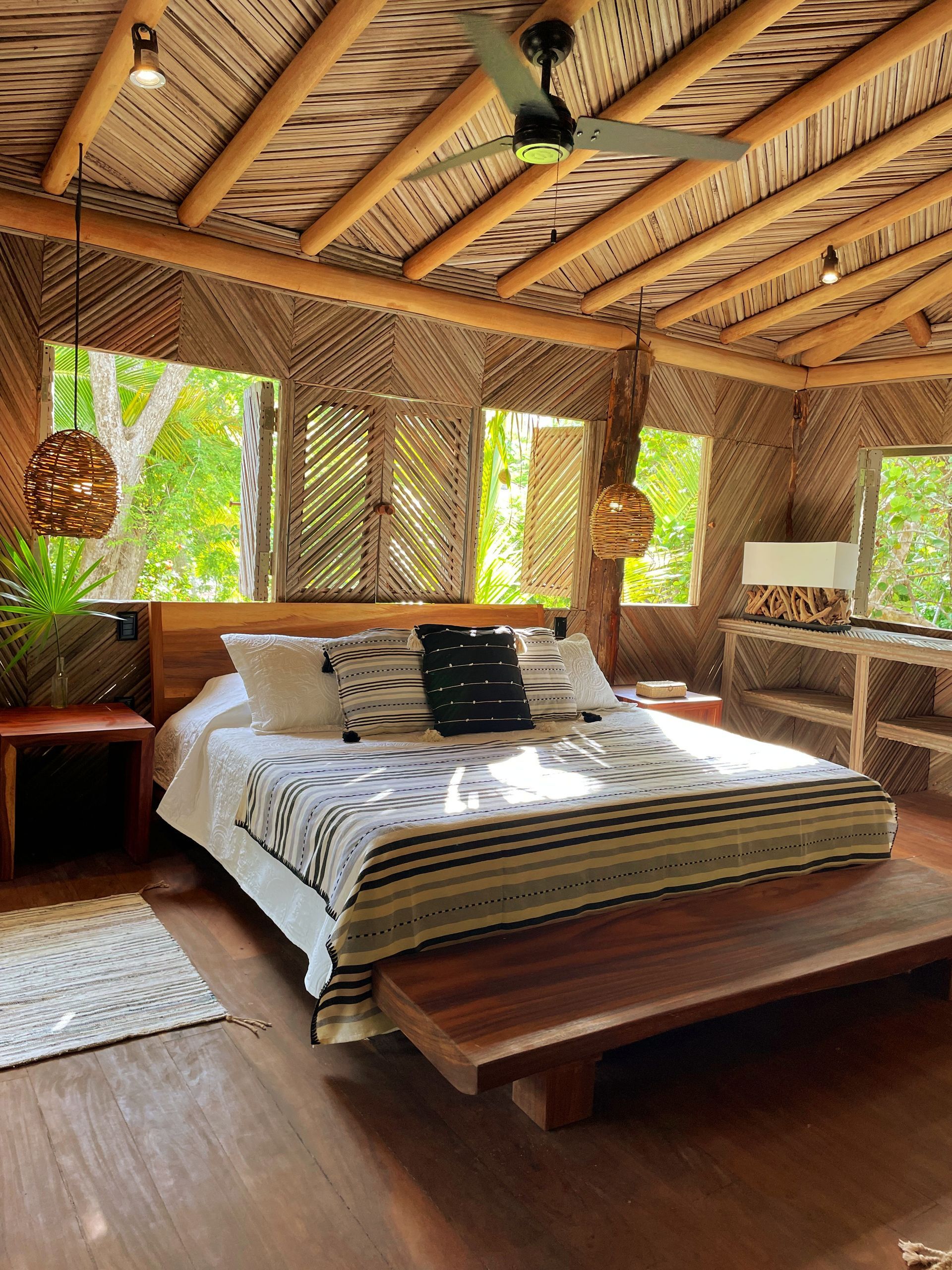 Dormitorio con cama de madera, detalles tejidos y techo de palma, con vista a los árboles.