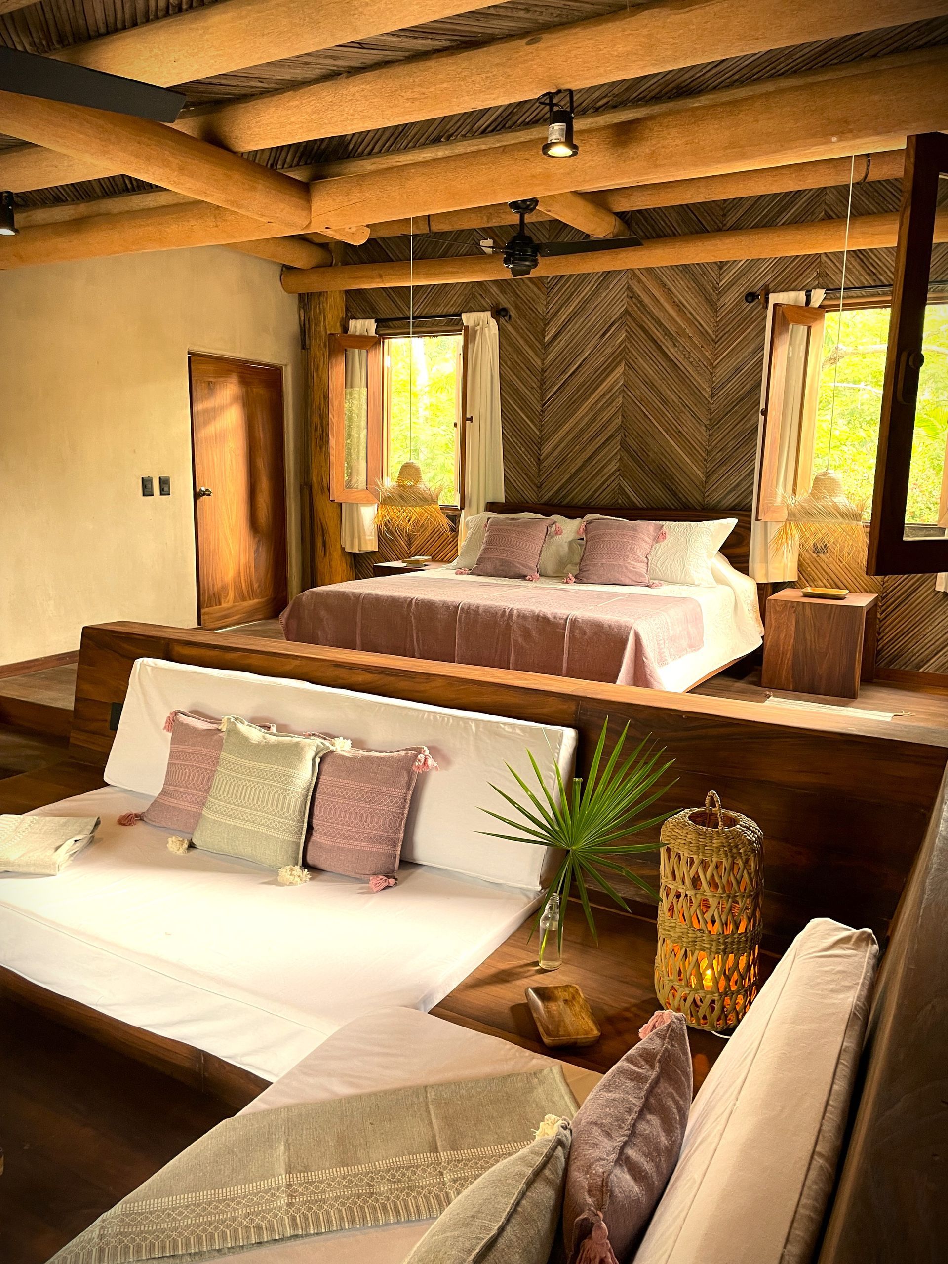 Dormitorio rústico con vigas de madera, cama con ropa de cama rosa, diván y luz natural.