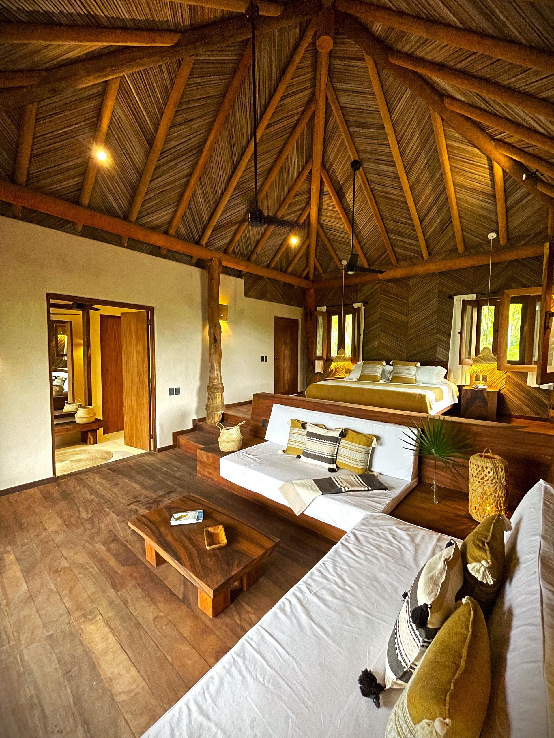 Lujoso dormitorio con techo alto de paja, cama tipo plataforma, sofás blancos, pisos de madera y entrada al baño.