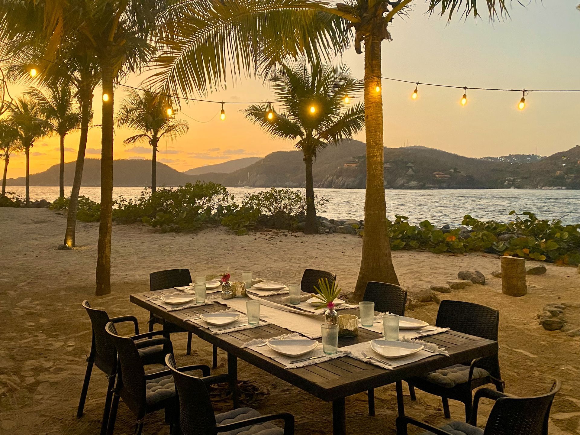 Mesa de comedor junto a la playa para una comida, palmeras, océano y puesta de sol.