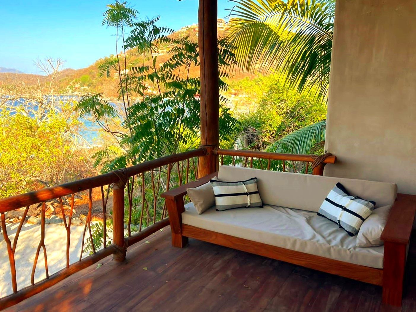 Porche de madera con sofá y vistas a la bahía, rodeado de vegetación. Ambiente soleado durante el día.