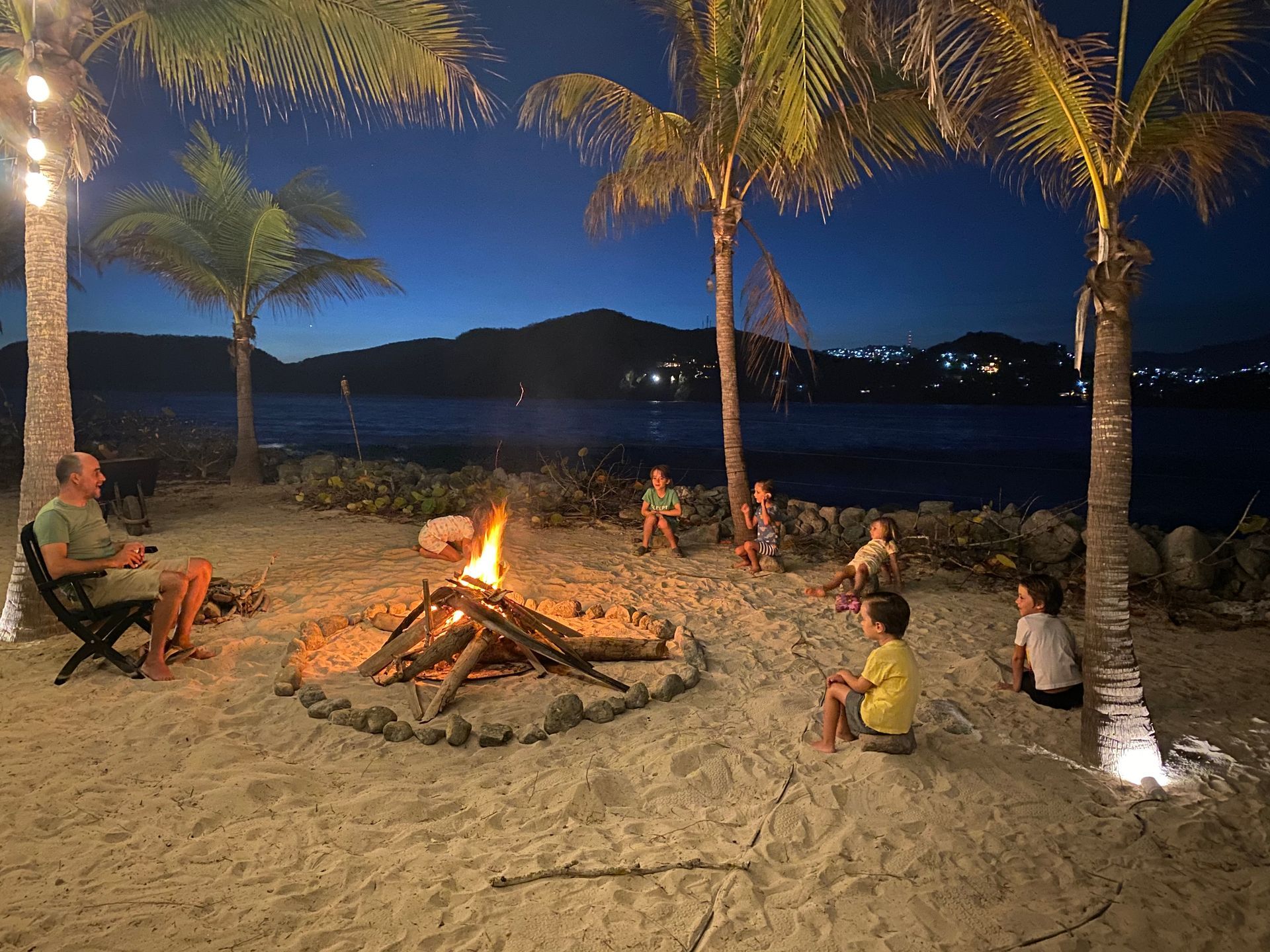Gente alrededor de una fogata en la playa bajo las palmeras por la noche.