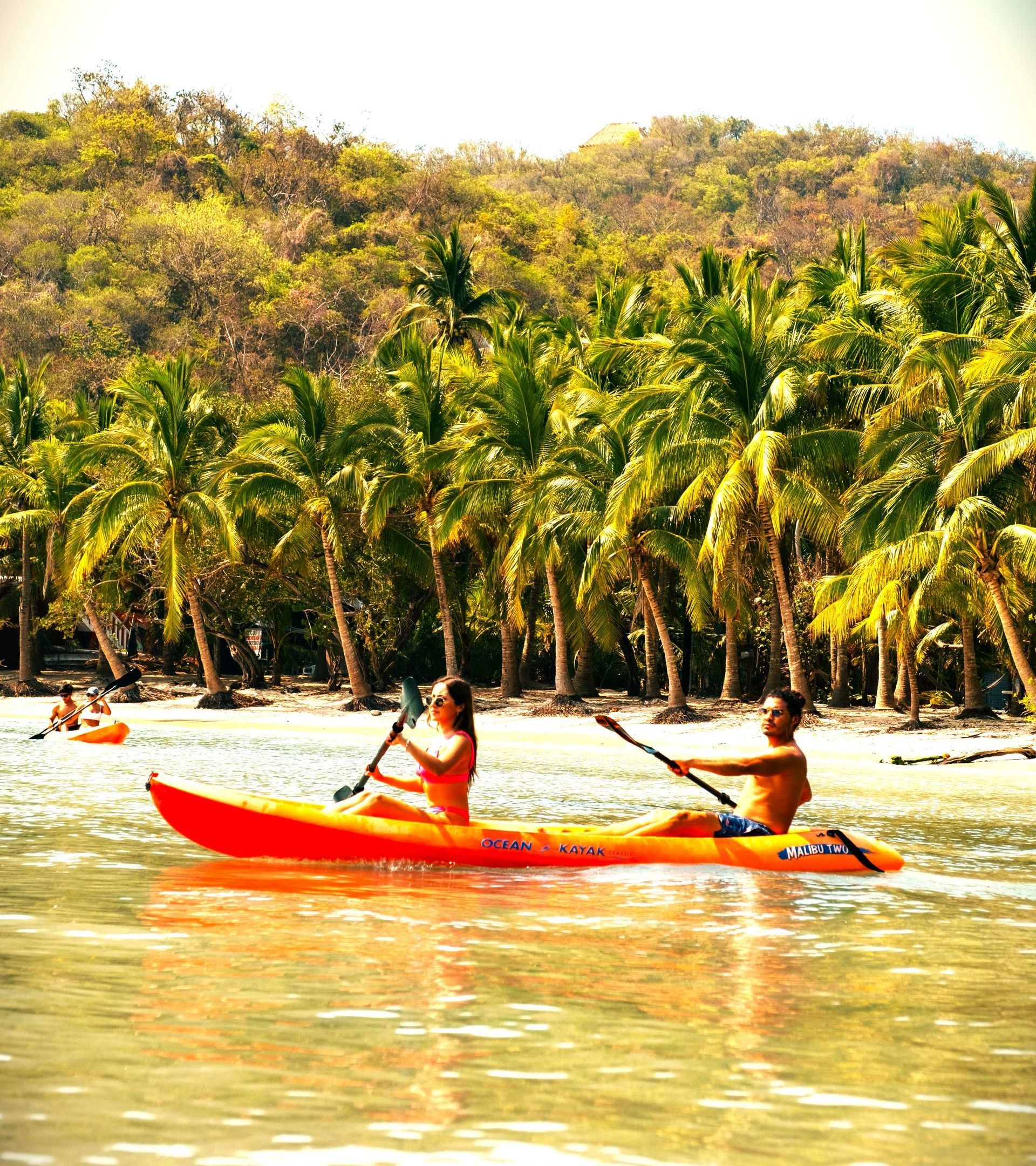 Dos personas navegando en un kayak naranja cerca de una playa tropical bordeada de palmeras.