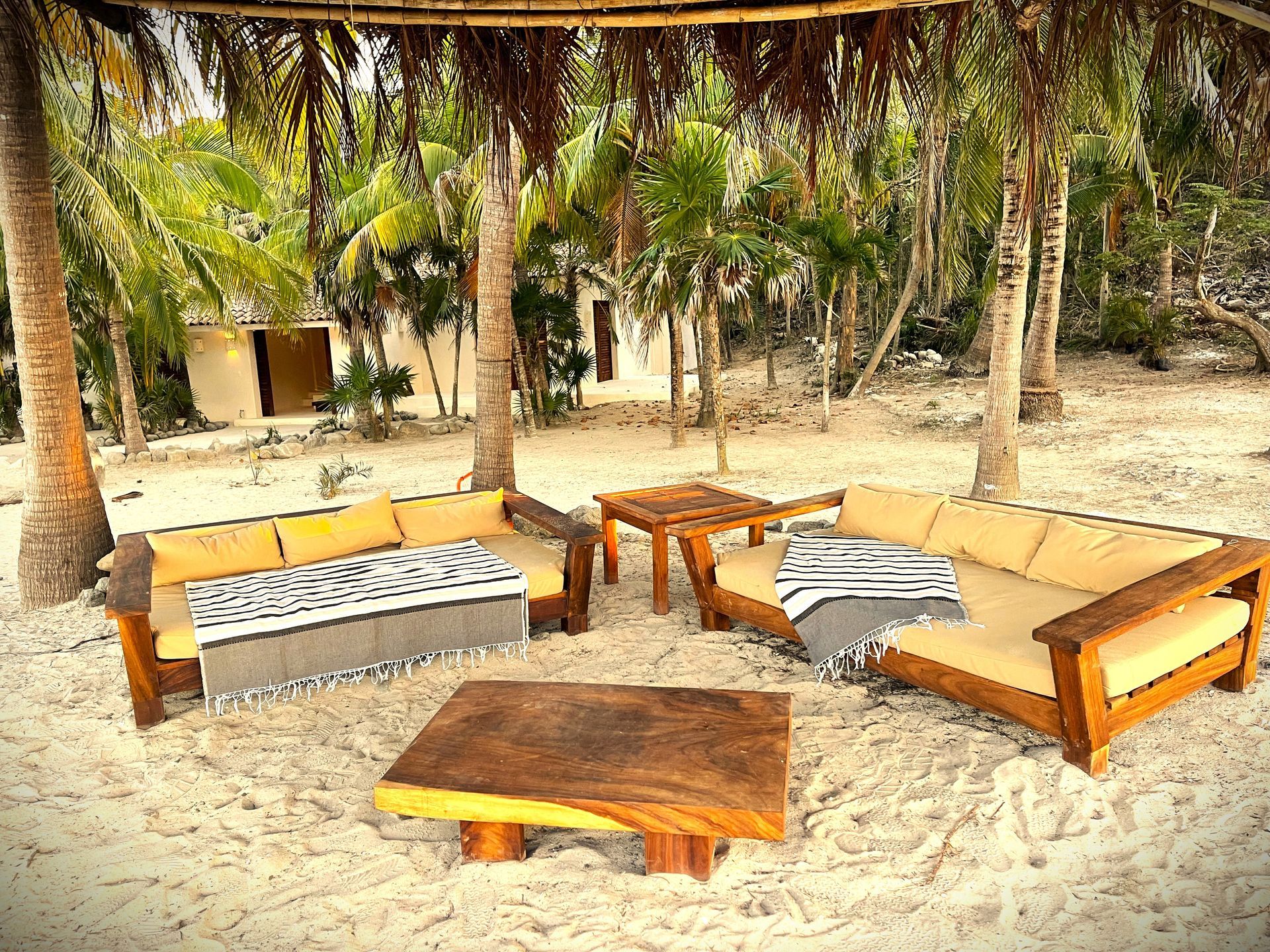 Zona de estar al aire libre en una playa de arena: sofás de madera, mesas de café y techo de paja.