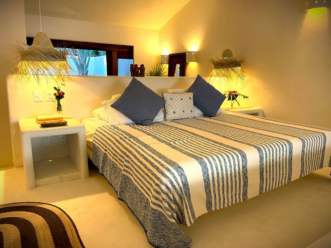 Dormitorio con colcha de rayas, almohadas azules, pantallas de paja y mesita de noche blanca.