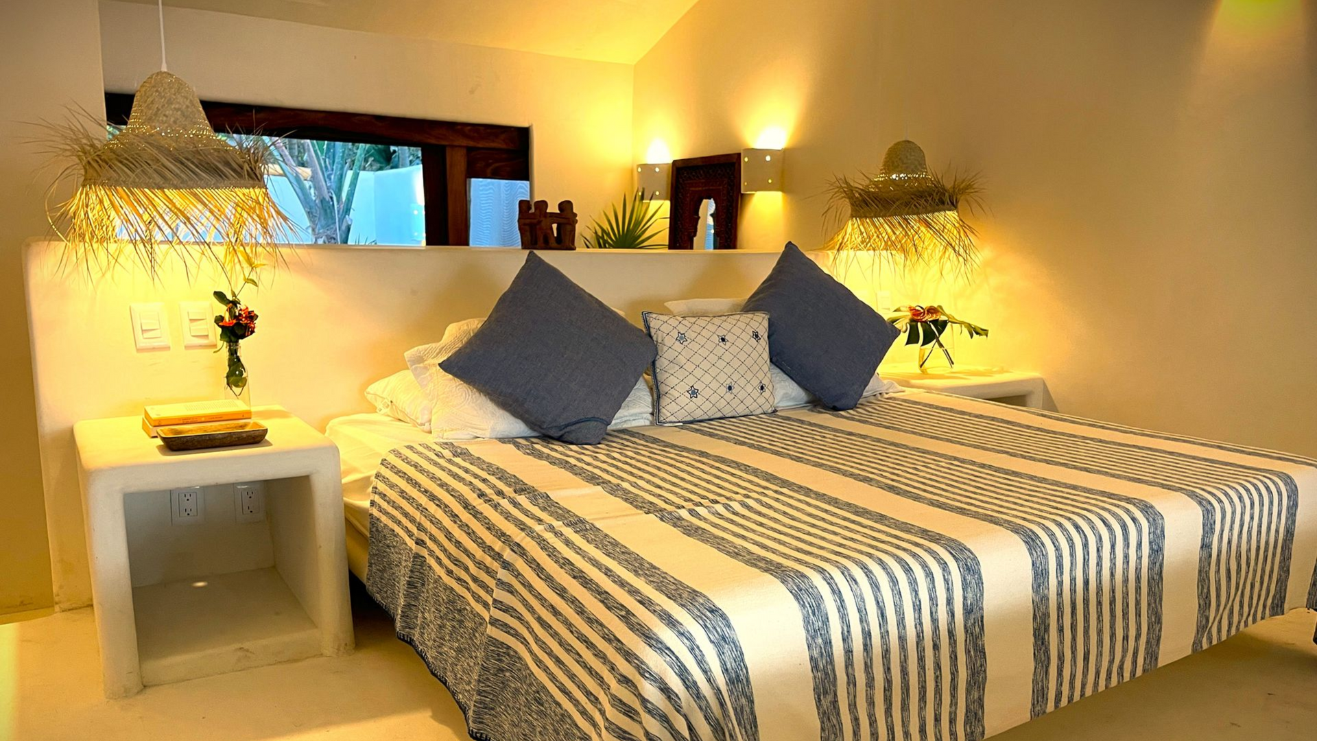 Dormitorio con colcha de rayas, almohadas azules, pantallas de paja y mesita de noche blanca.