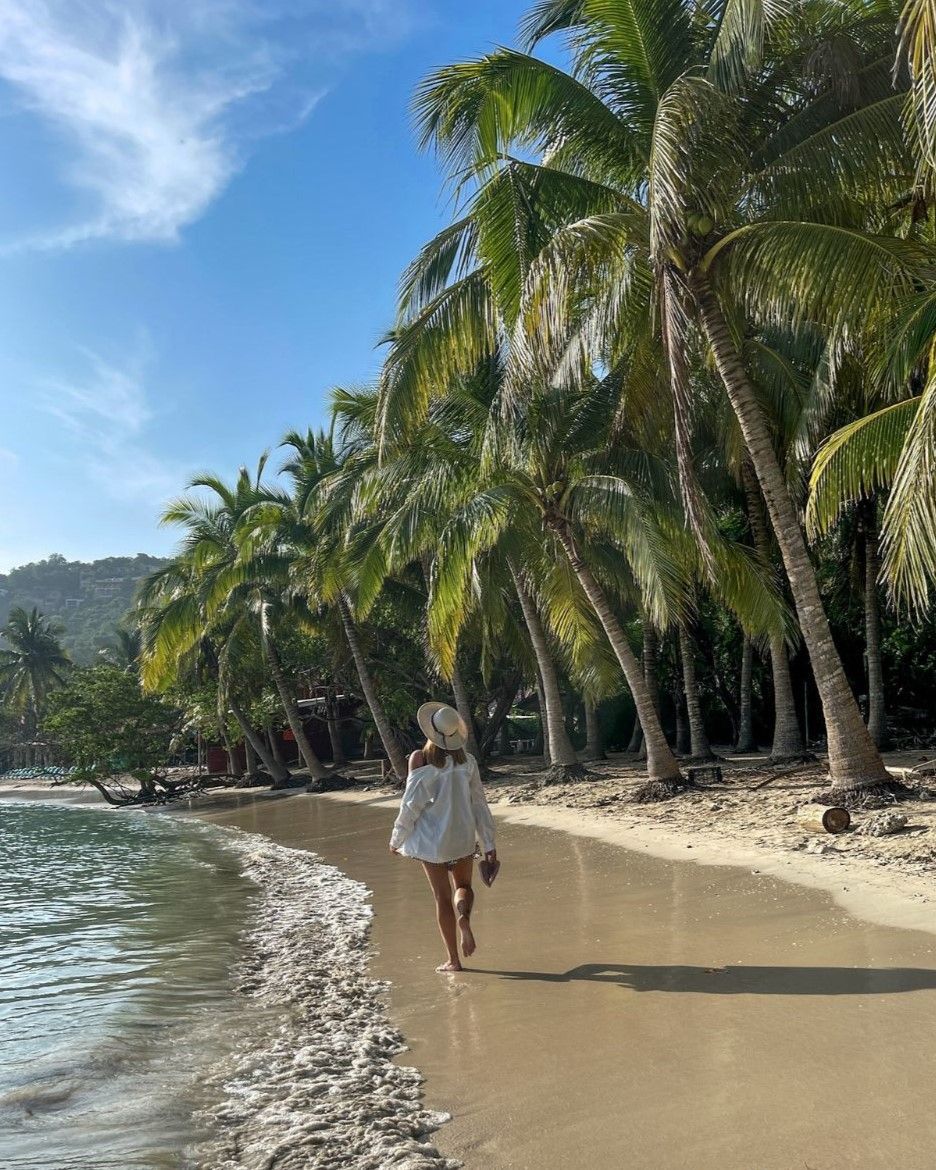 Mujer caminando por una playa, con palmeras y océano en un día soleado.