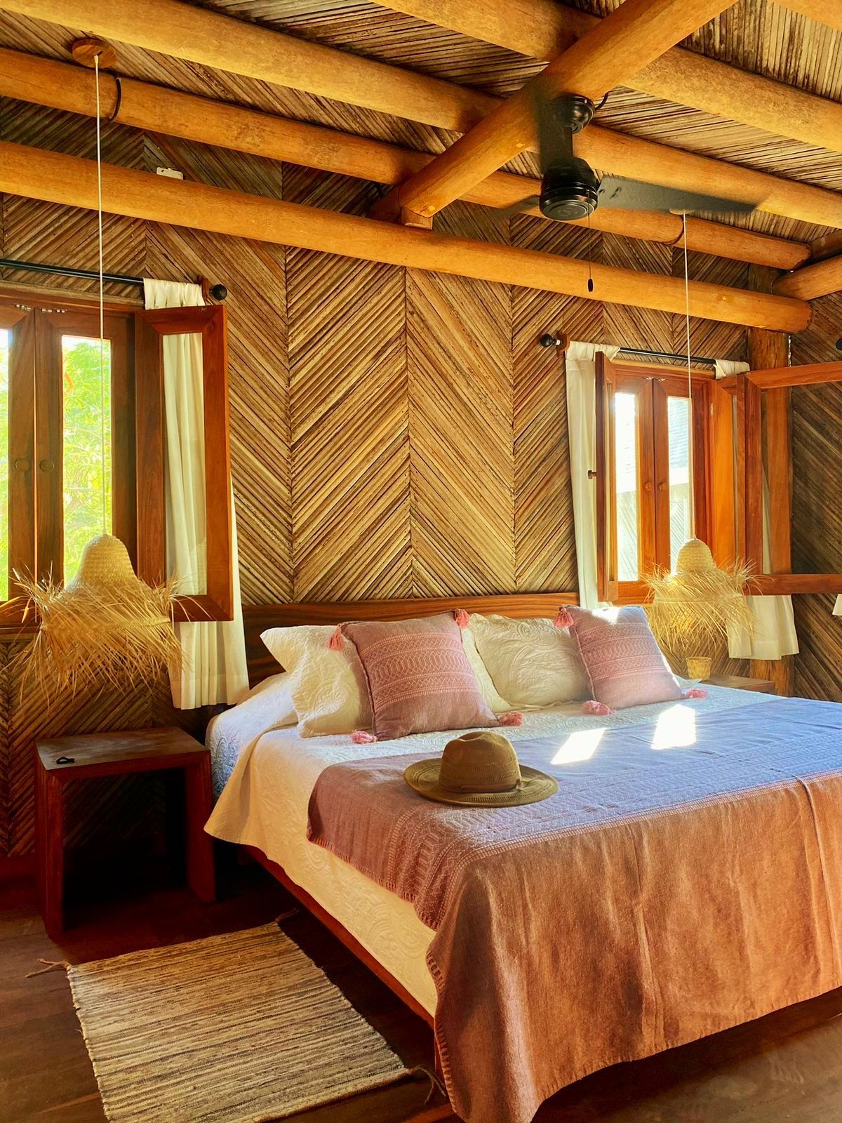 Acogedor dormitorio con paredes de madera tejida, cama y mantas rosas, pantallas de lámparas de paja y un sombrero.