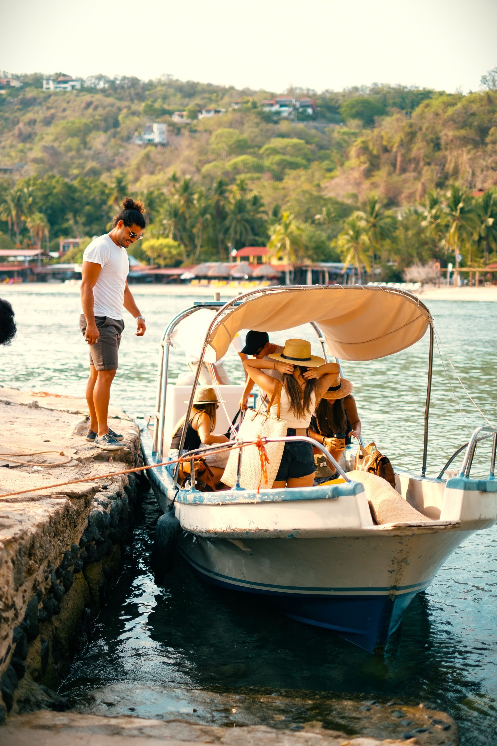 Hombre ayudando a la gente a subir a un barco en un muelle; día soleado, colinas verdes en el fondo.