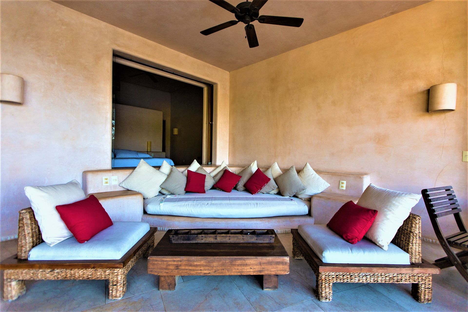 Zona de estar en el patio con paredes beige, muebles de madera y almohadas de color rojo/crema.