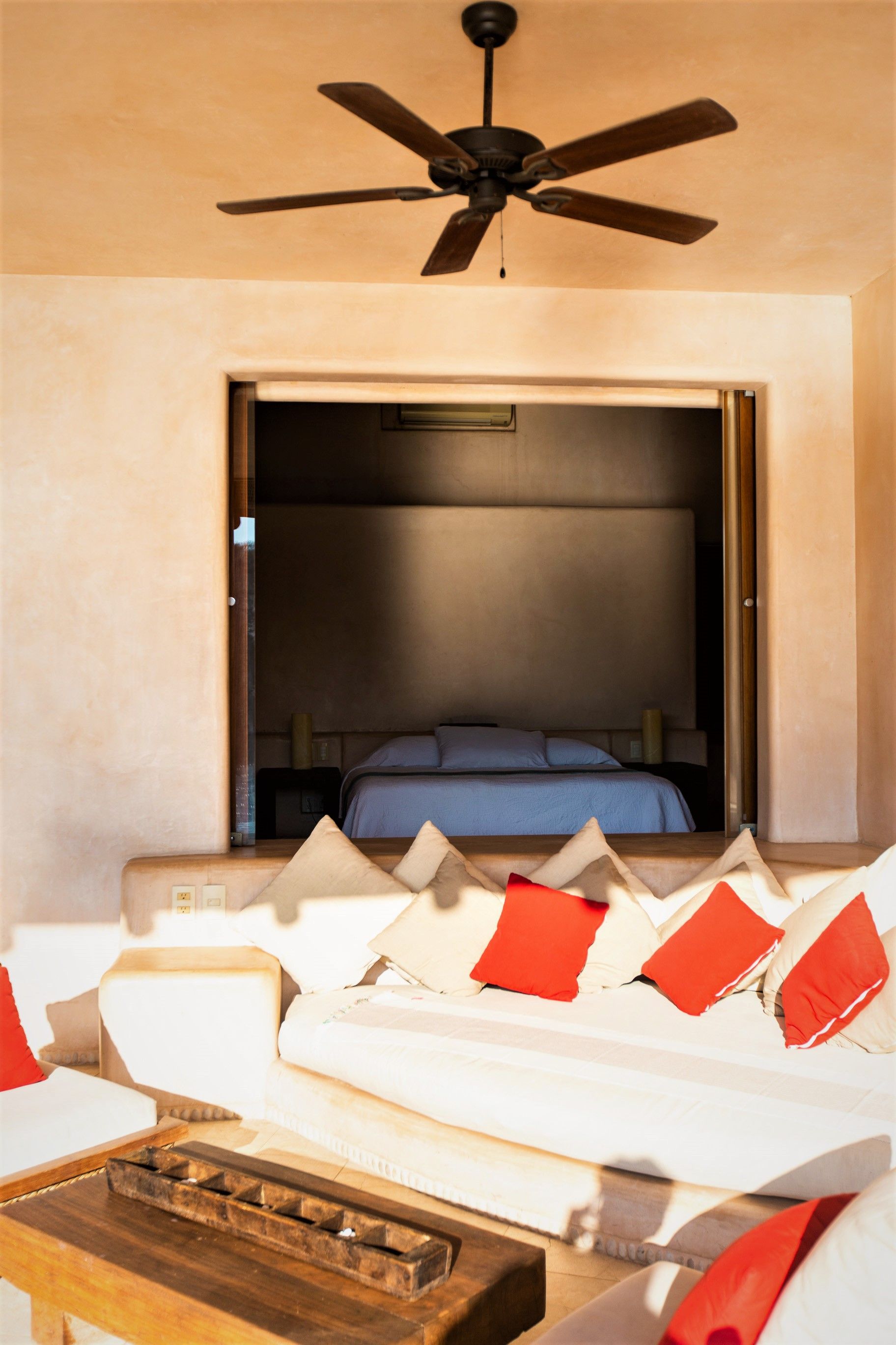 Zona de estar al aire libre con vista al dormitorio, cojines blancos con almohadas rojas, ventilador de techo.