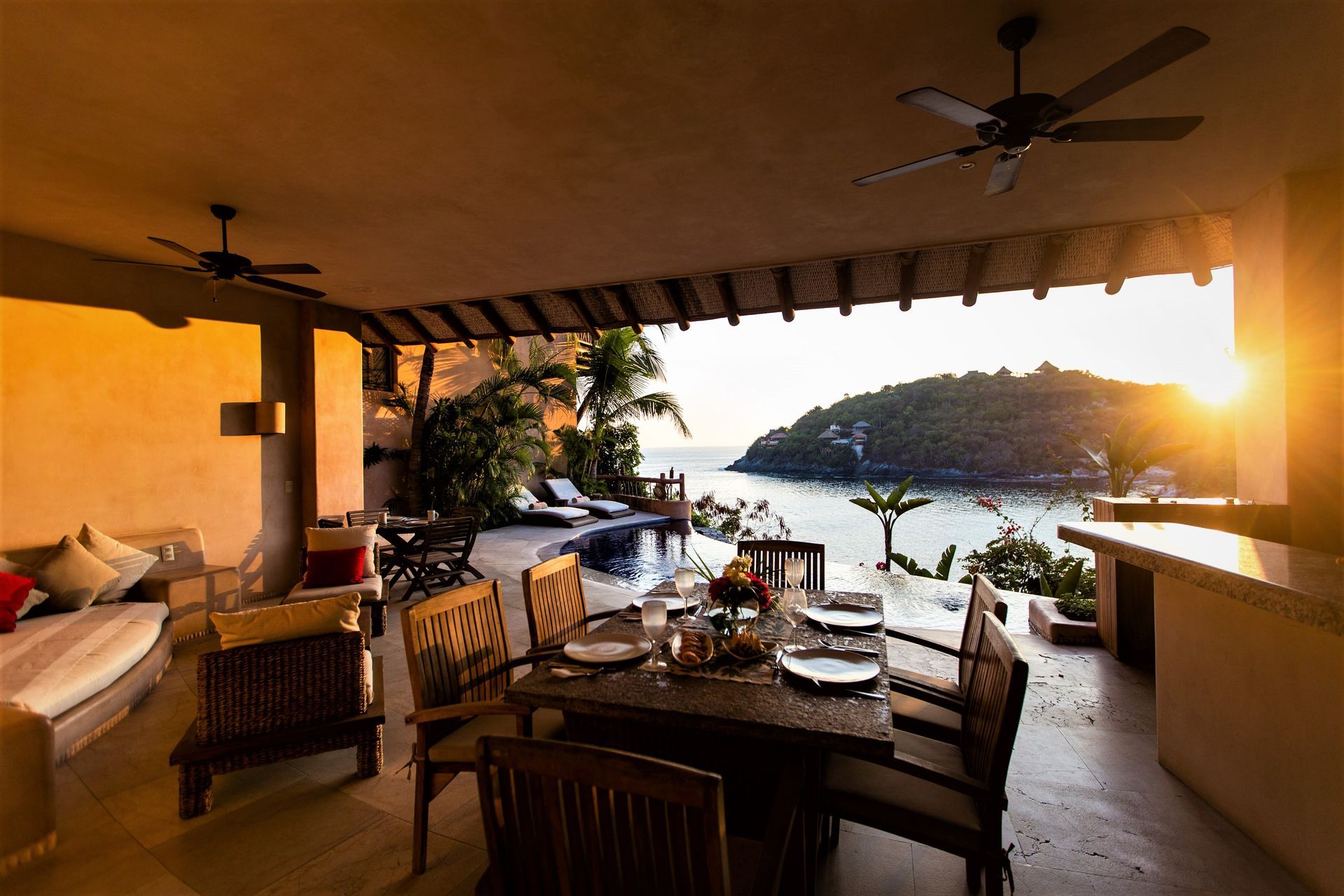 Comedor al aire libre con vista al mar al atardecer. Mesa de madera, sillas y piscina.