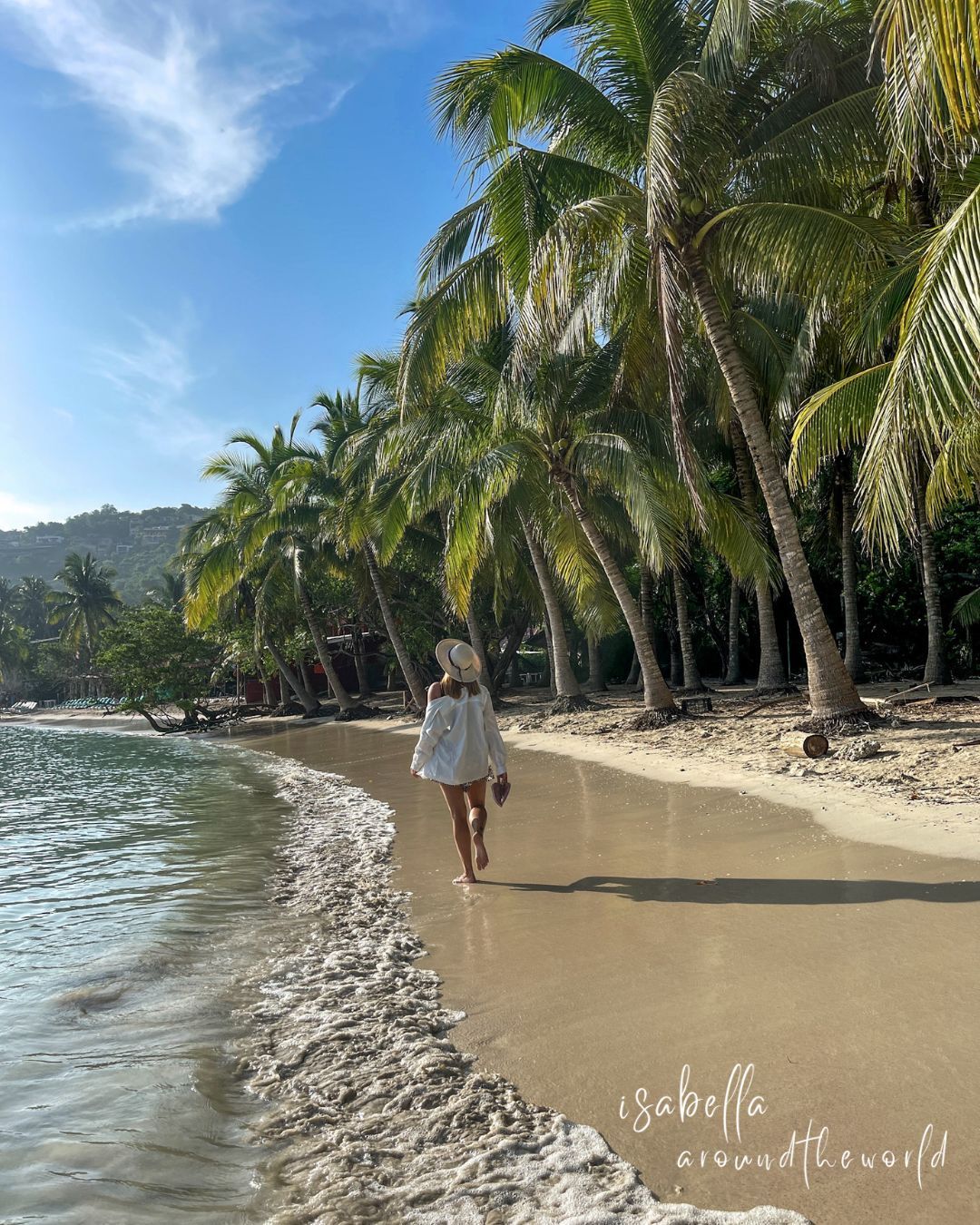 Mujer caminando por una playa soleada, enmarcada por palmeras y el océano, con un vestido blanco.