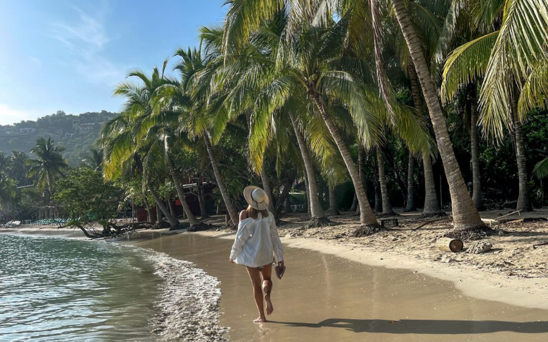Mujer caminando por una playa soleada, enmarcada por palmeras y el océano, con un vestido blanco.