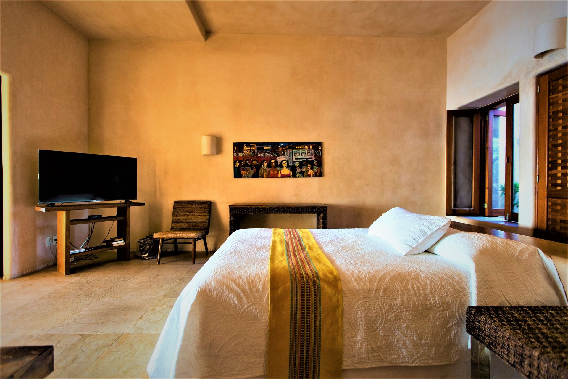 Dormitorio con cama, televisor y ventana. Paredes color crema y suelo de baldosas claras.