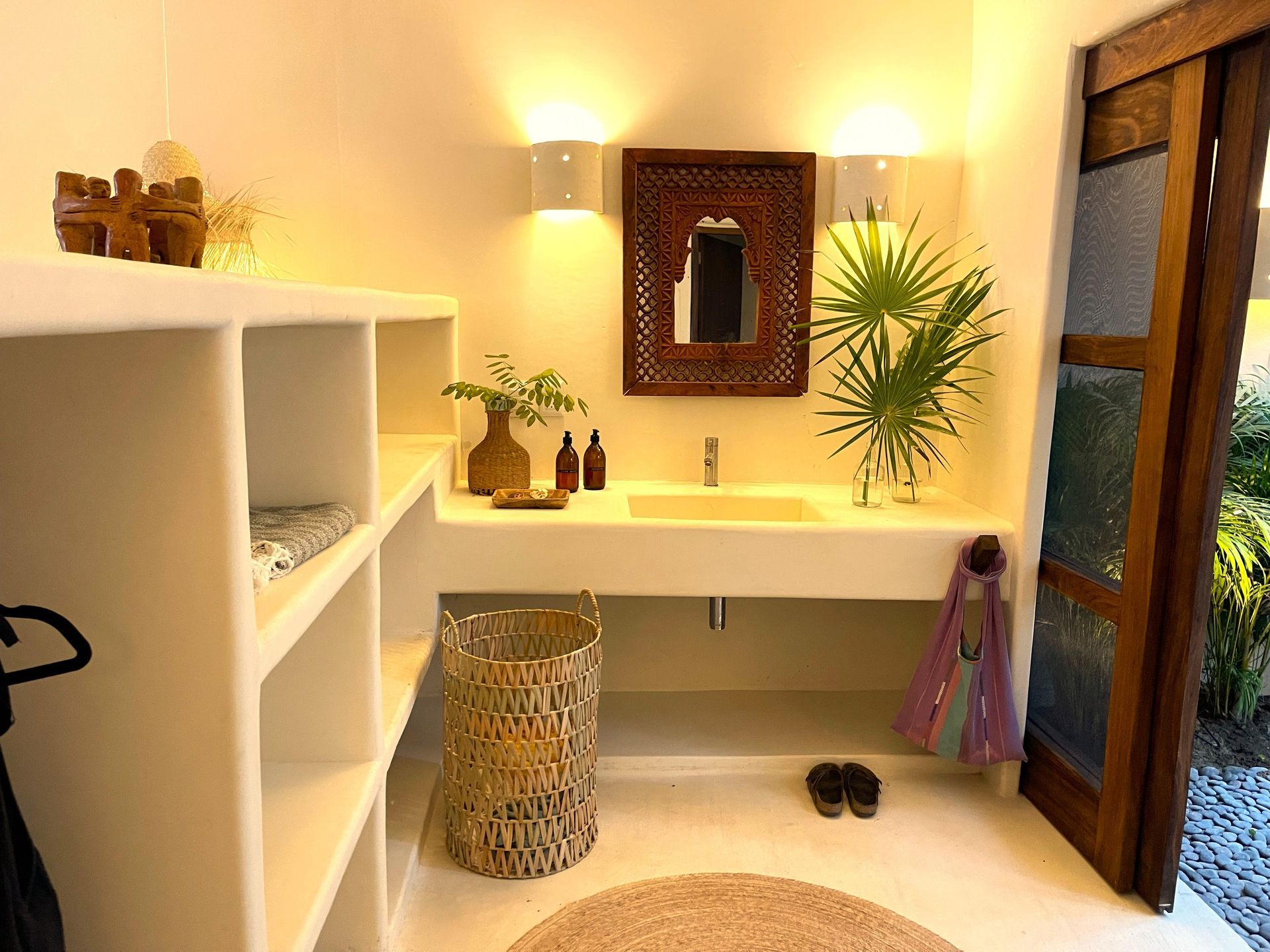 Baño luminoso con estanterías blancas, lavabo, espejo de madera y puerta abierta a la vegetación.