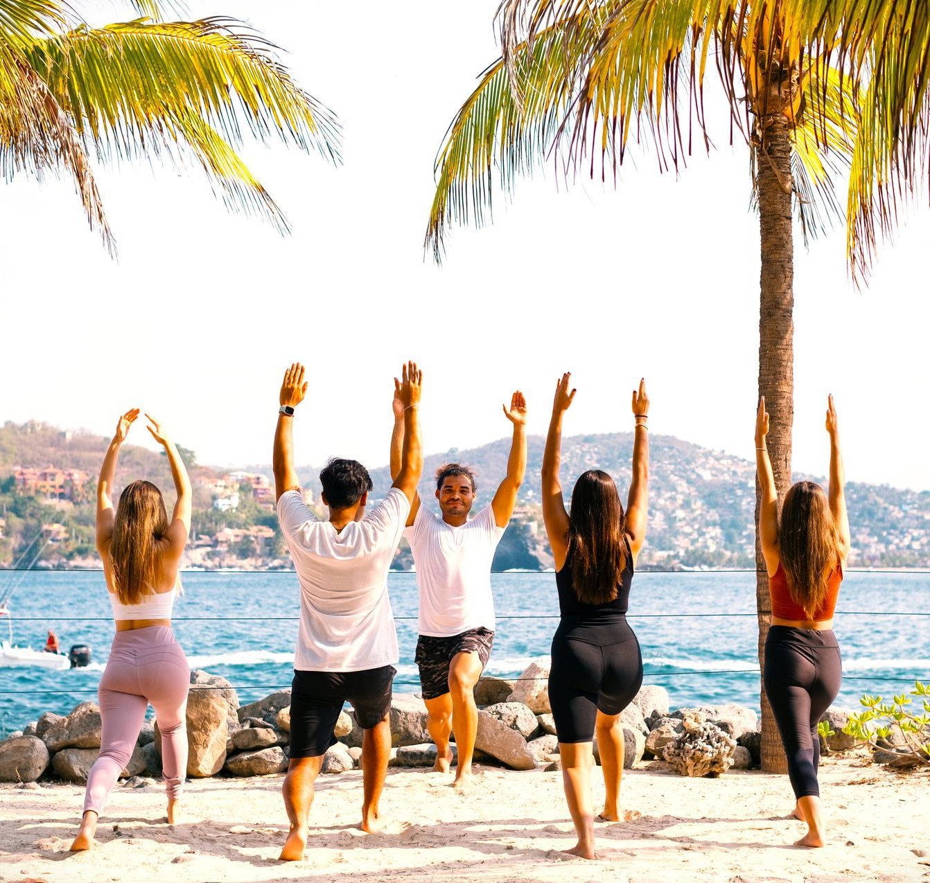 Personas practicando yoga en la playa bajo palmeras, con los brazos en alto. Cielo azul despejado.