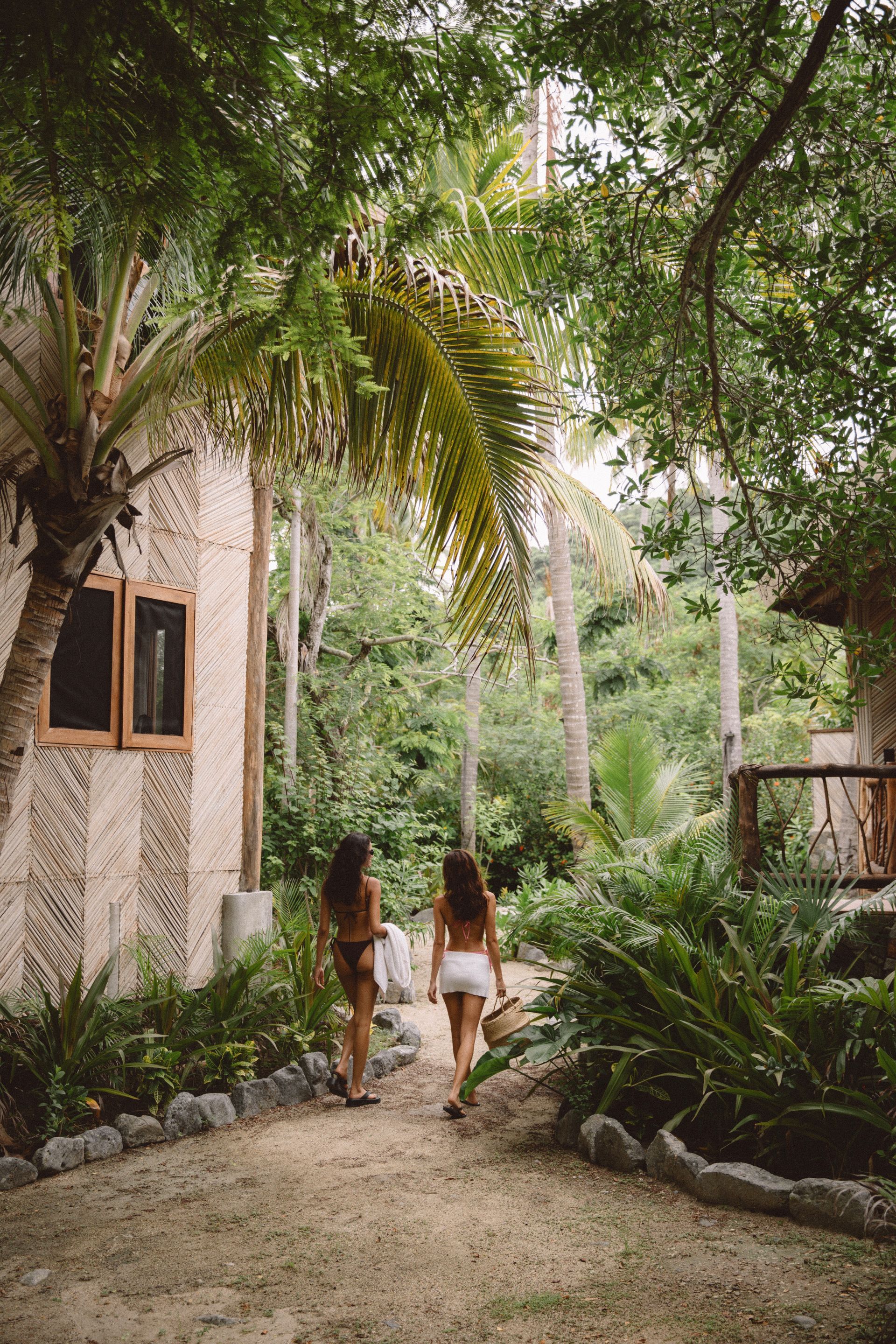 Dos mujeres caminando por un sendero de arena a través de un exuberante follaje tropical, hacia un edificio.