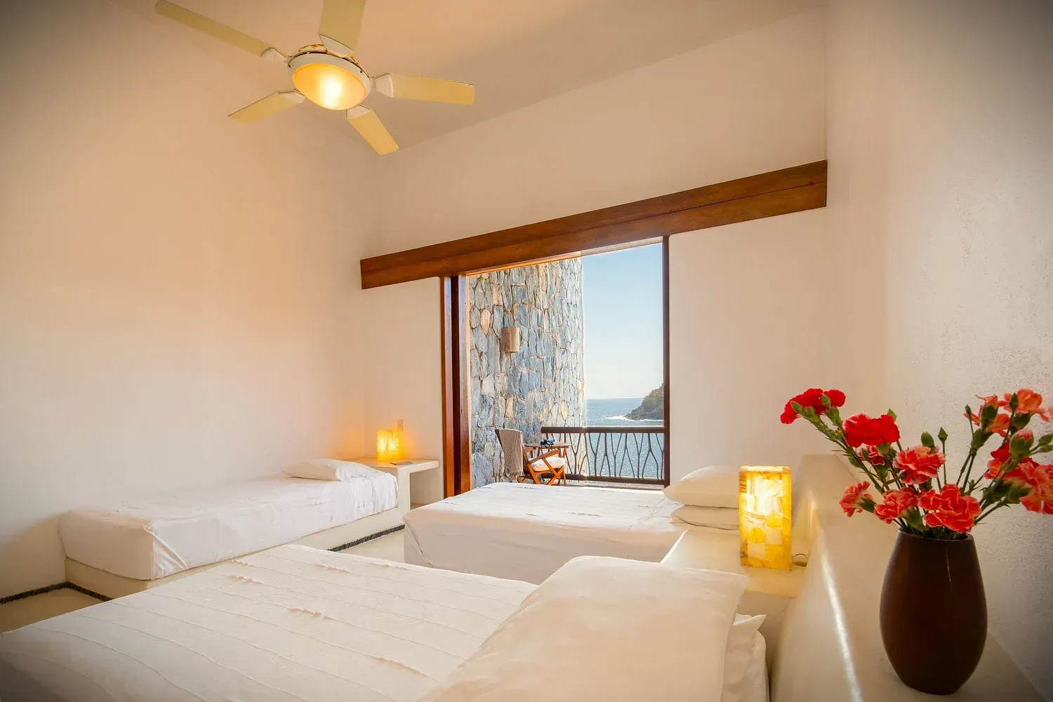 Dormitorio con tres camas, paredes blancas, vista al mar, jarrón con flores y velas encendidas.
