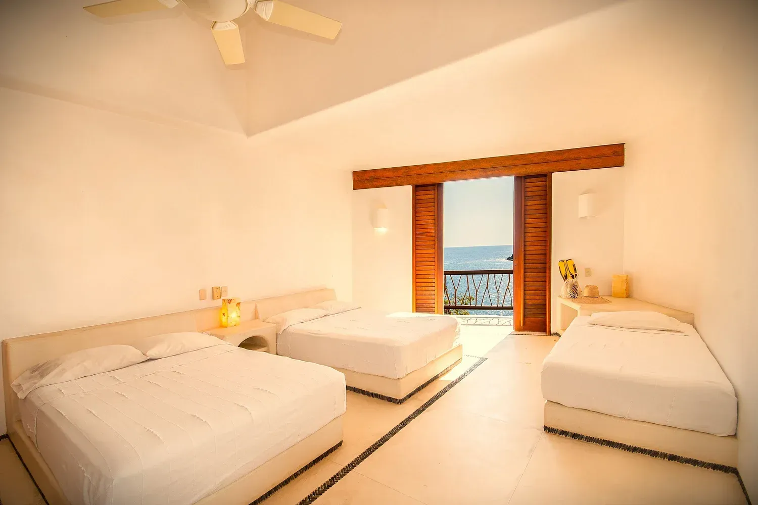Amplia habitación de hotel con tres camas blancas, puertas abiertas a un balcón y paredes claras.