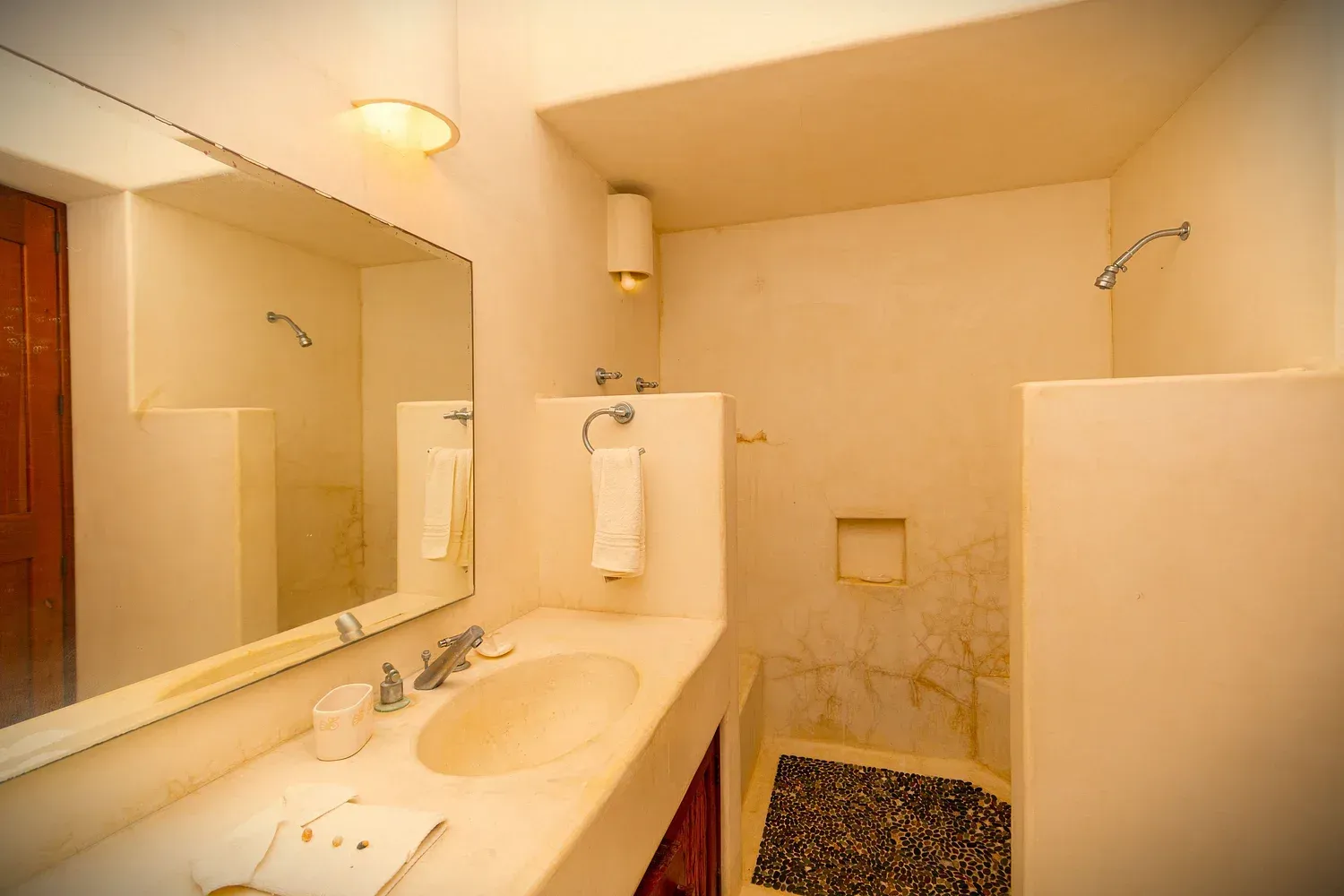 Baño con paredes en blanco, lavabo, espejo y ducha. Hay una toalla colgada en la pared y una pequeña alfombra en la ducha.