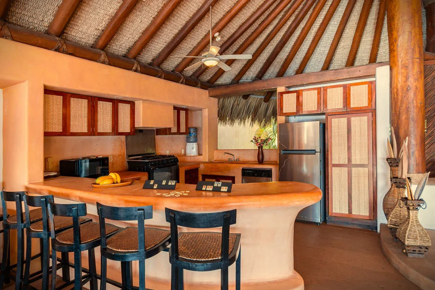 Cocina con techo de paja, paredes anaranjadas, gabinetes de madera y barra con taburetes.