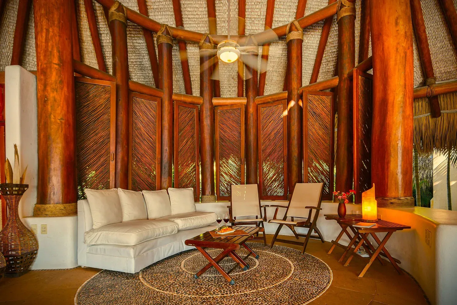 Una sala de estar redonda de estilo tropical con vigas de madera, paredes tejidas, muebles blancos y ventilador de techo.