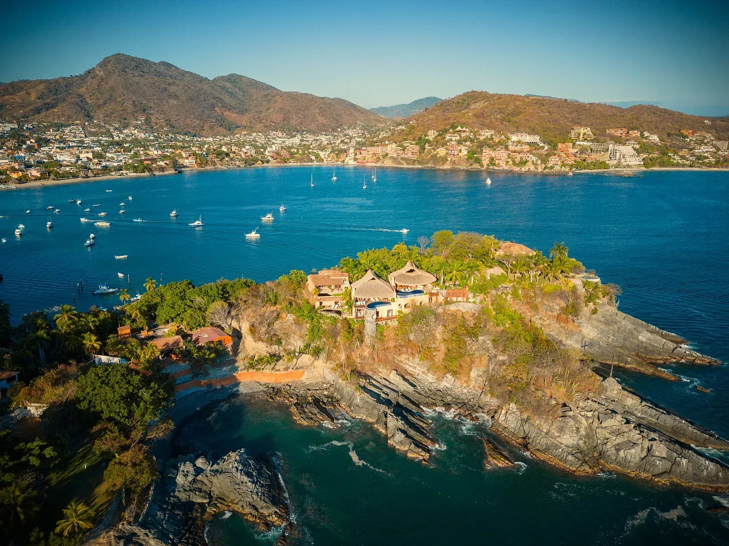 Isla con edificios, rodeada de agua azul y montañas al fondo.