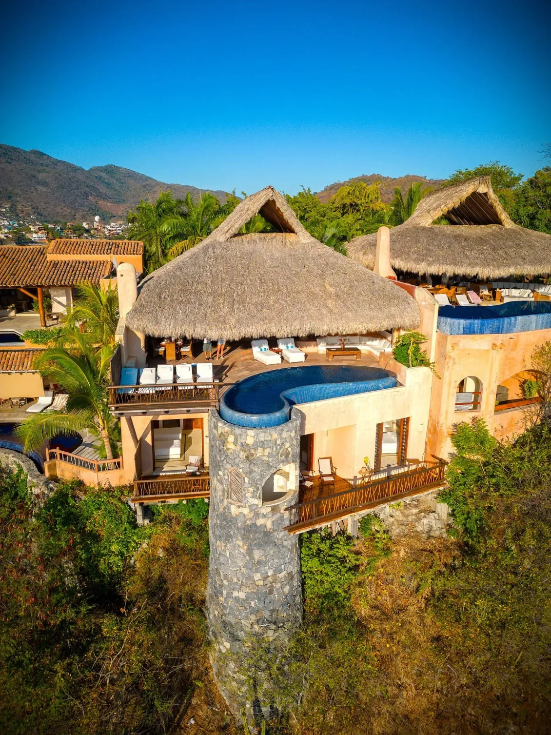 Villa de lujo con techos de paja, piscina infinita azul, vista al mar, ubicada en un exuberante entorno verde.