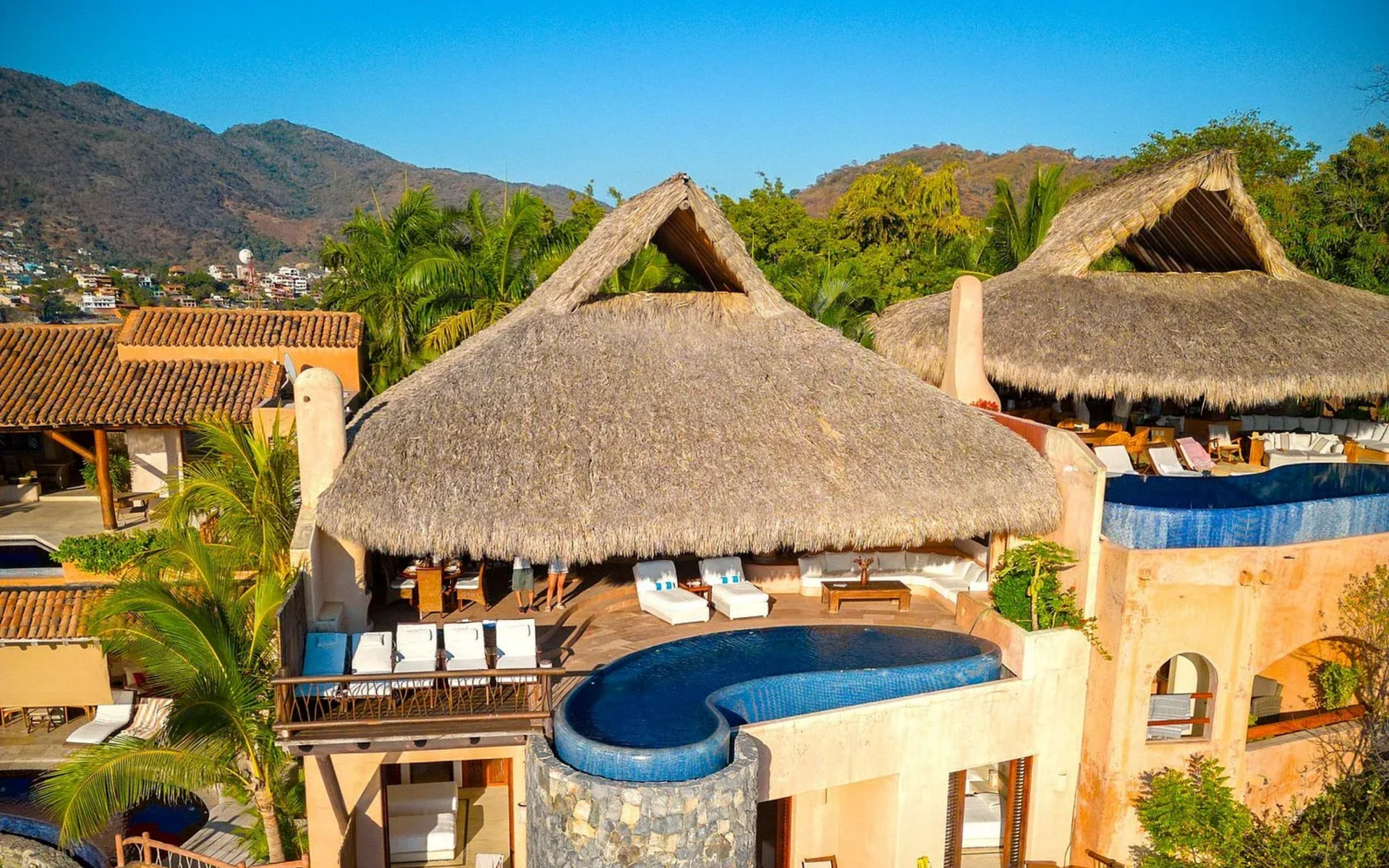 Villa de lujo con techos de paja, piscina infinita azul, vista al mar, ubicada en un exuberante entorno verde.
