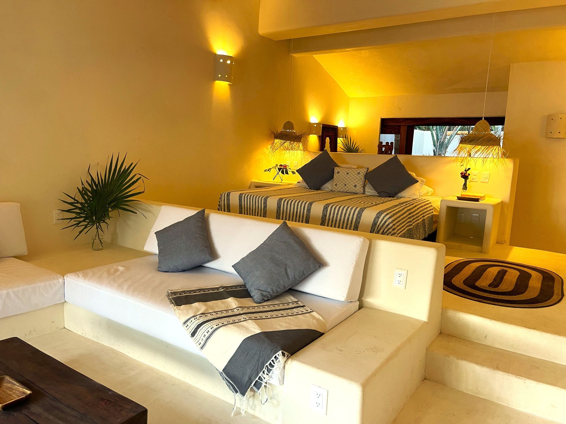 Dormitorio blanco y amarillo con cama elevada, sofá e iluminación cálida.