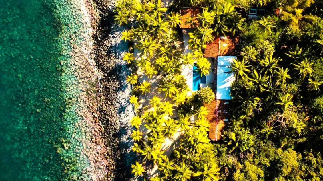 Vista aérea: agua turquesa, orilla rocosa, exuberantes árboles verdes que rodean un edificio con una piscina azul.